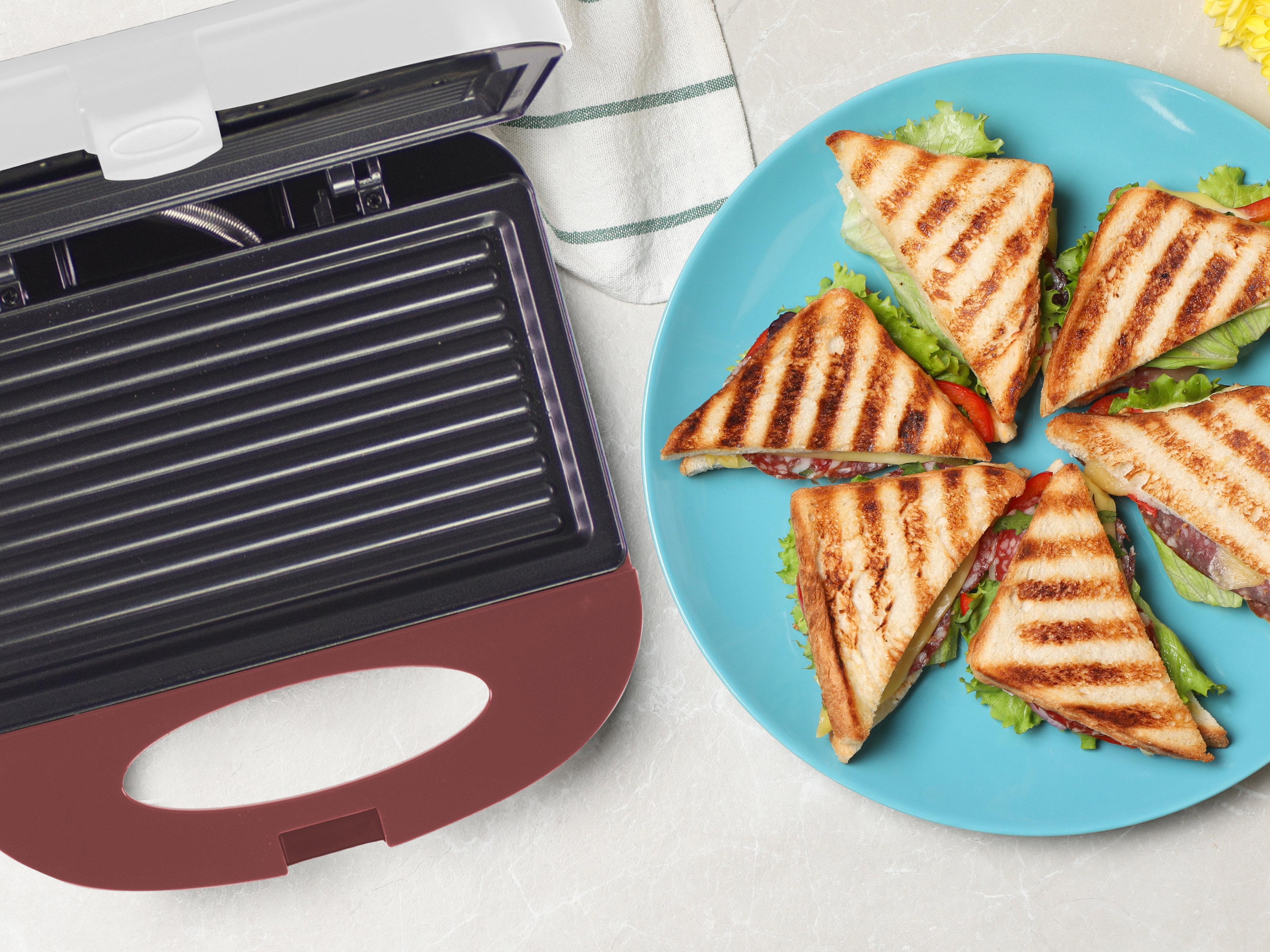 Griglia aperta con panini su piatto blu. I panini hanno lattuga, formaggio e salame. Panno bianco e fiore giallo visibili.