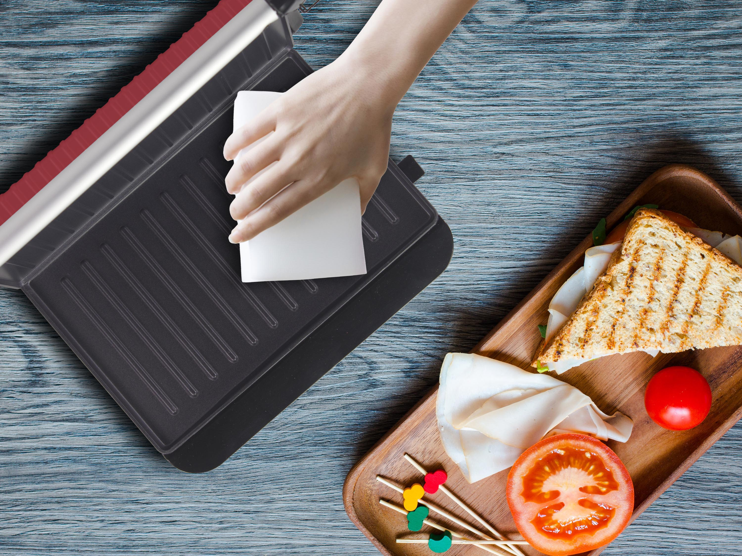 Una mano pulisce una griglia. Un sandwich, pomodoro e spiedini su un vassoio di legno. Sfondo di legno blu.