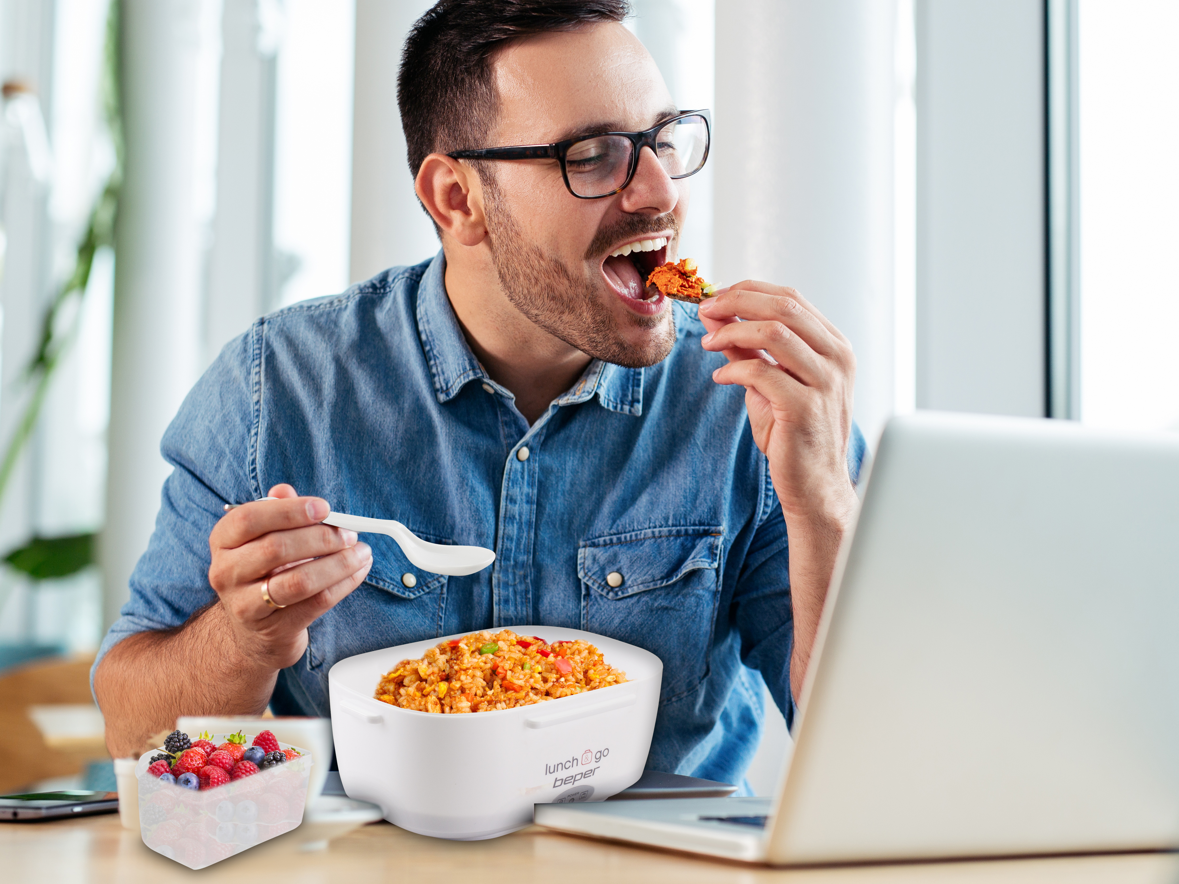 Uomo che mangia cibo da un contenitore bianco, con un cucchiaio, con un laptop davanti. Un contenitore di frutta a sinistra.