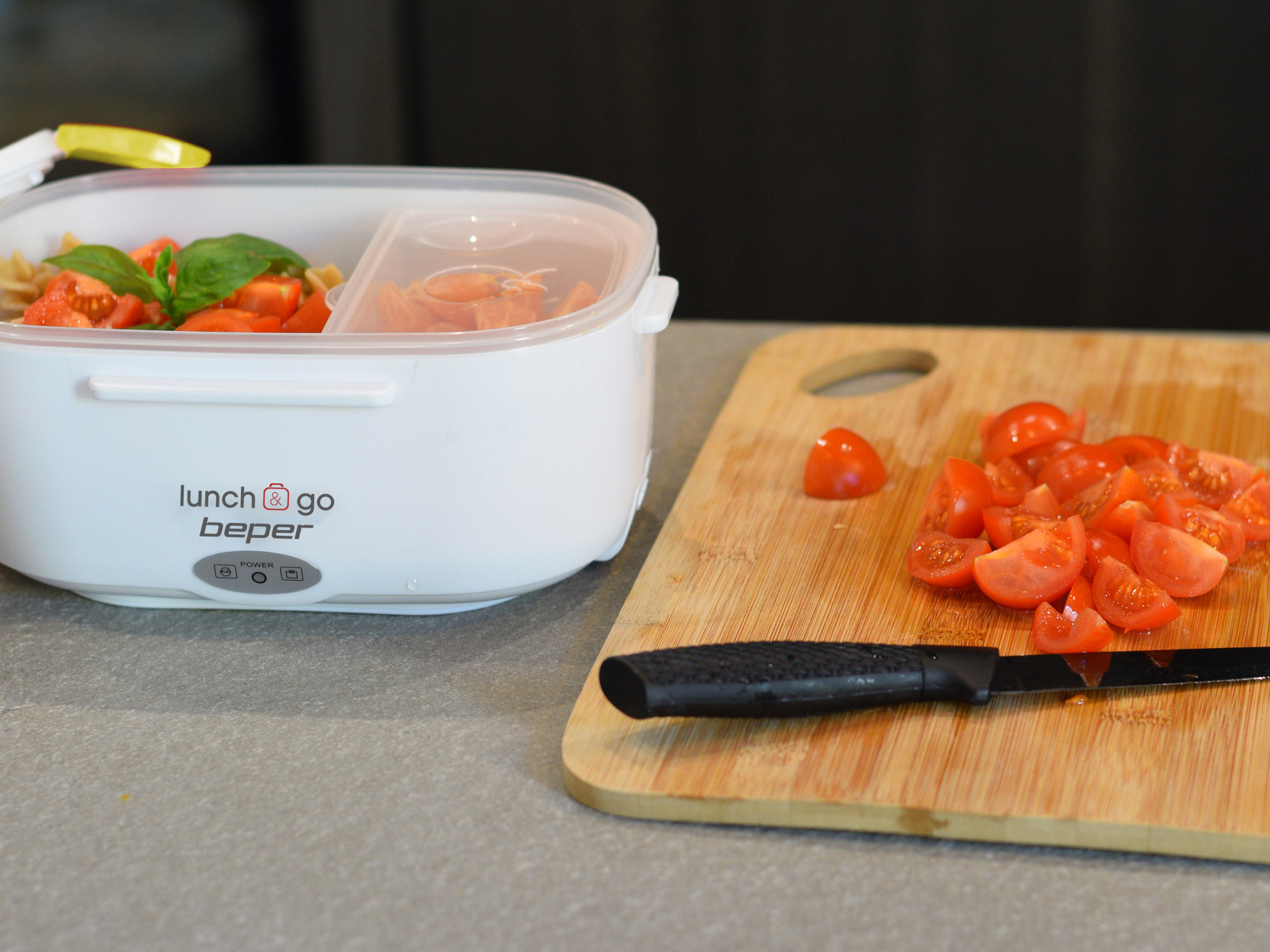 Un portapranzo bianco con cibo, pomodori su un tagliere e un coltello.