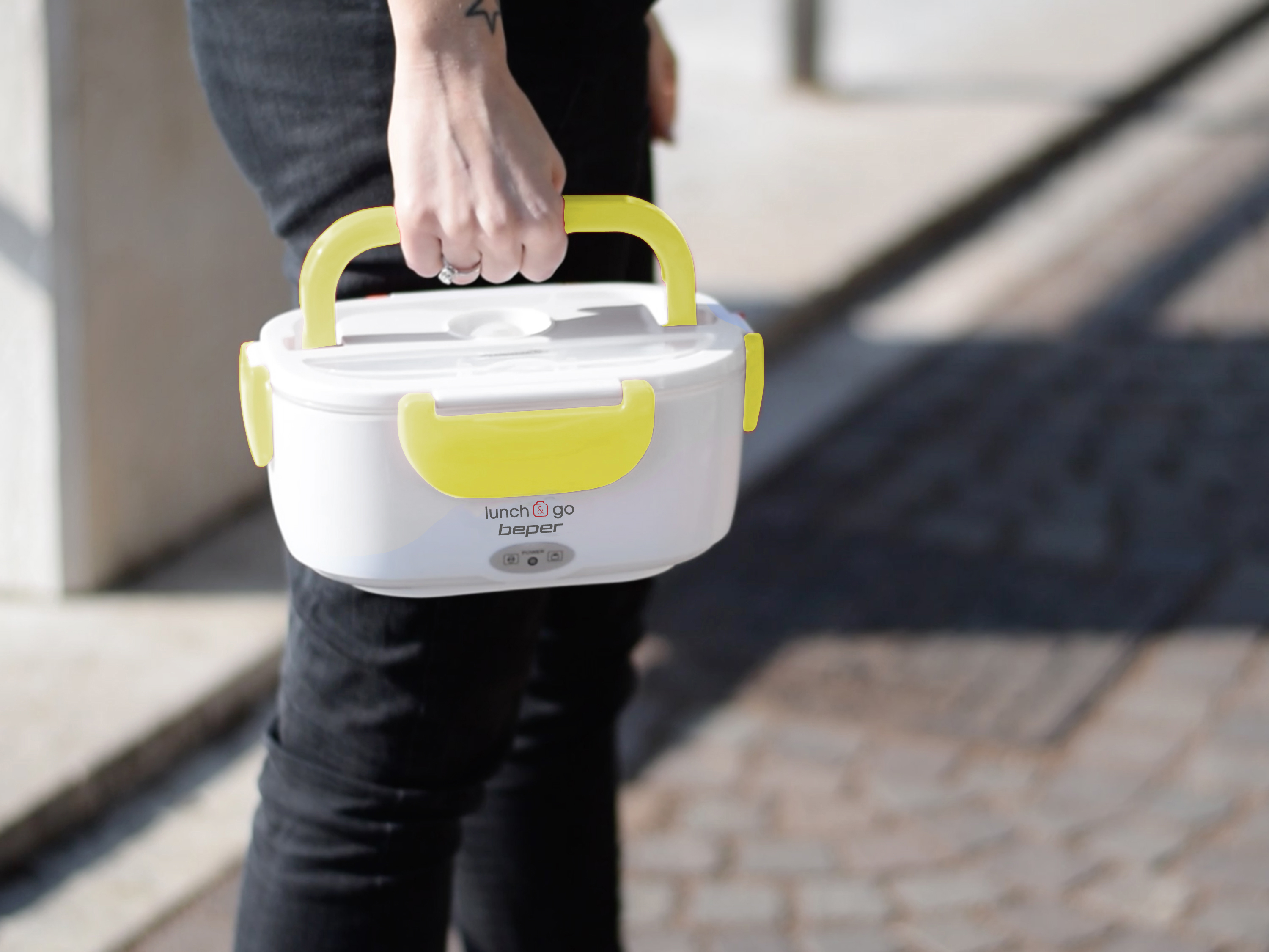 Una persona tiene in mano un portapranzo bianco con manici gialli. La scatola ha il testo 'lunch & go beper'. La persona indossa jeans neri.