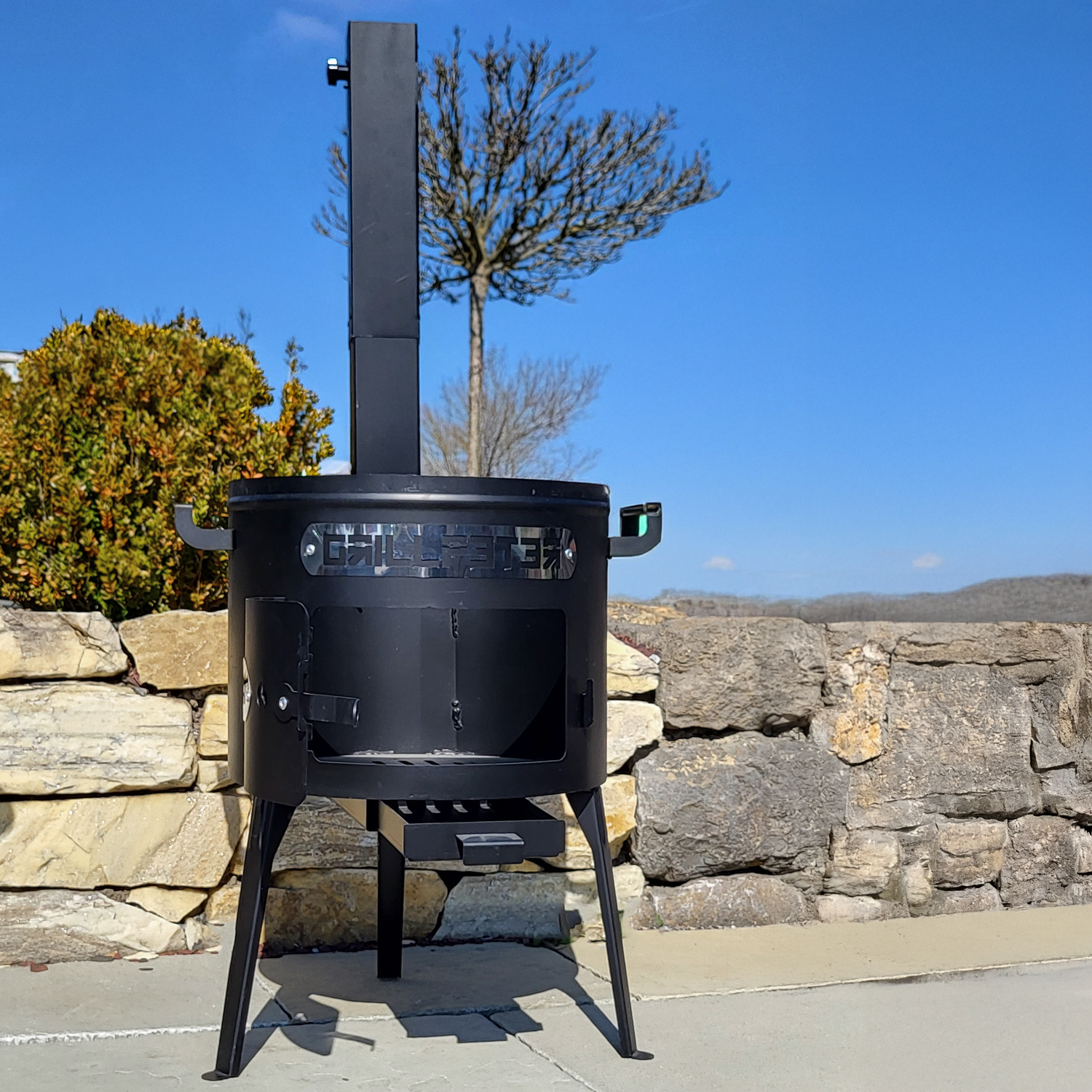 Ein schwarzer Outdoor-Kocher steht auf Beton. Im Hintergrund befindet sich eine Steinmauer mit blauem Himmel. Bäume und Büsche sind ebenfalls sichtbar.