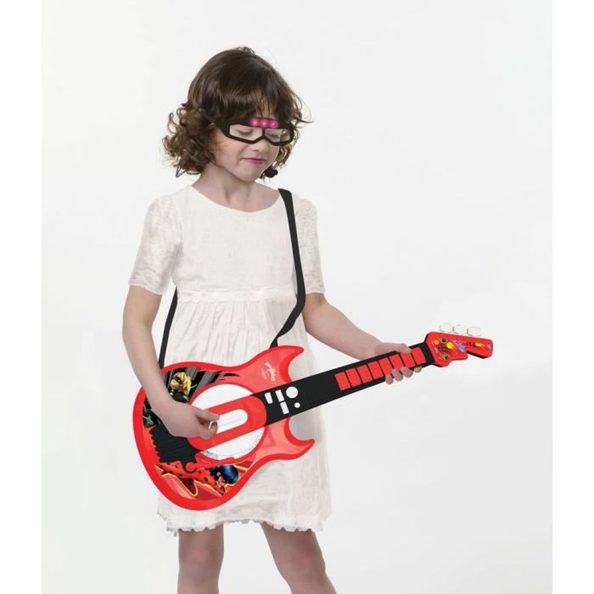Una niña con vestido blanco sostiene una guitarra de juguete roja. Lleva gafas con luces. Una correa negra alrededor del cuello.