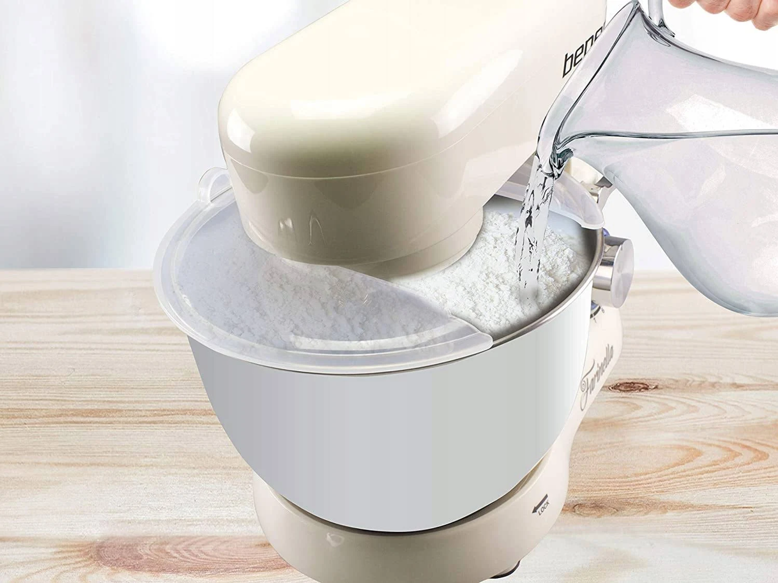 Un mixer con farina nella ciotola viene riempito con acqua da una brocca. Il mixer è bianco e beige.