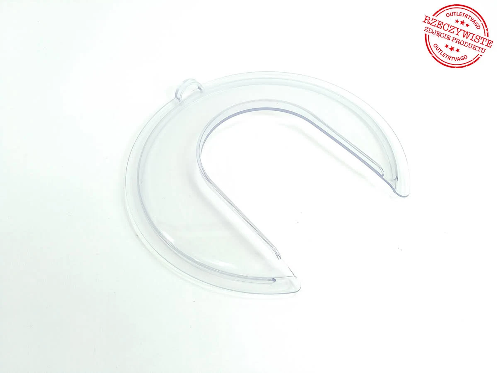 Oggetto di plastica trasparente a forma di mezzaluna, probabilmente un componente di un elettrodomestico da cucina, su una superficie bianca. Timbro rosso in alto a destra.