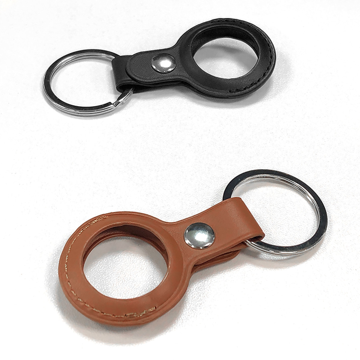 Dwa breloczki na białym tle. Jeden czarny, jeden brązowy. Każdy ma pierścień i skórzany uchwyt z metalowym guzikiem.