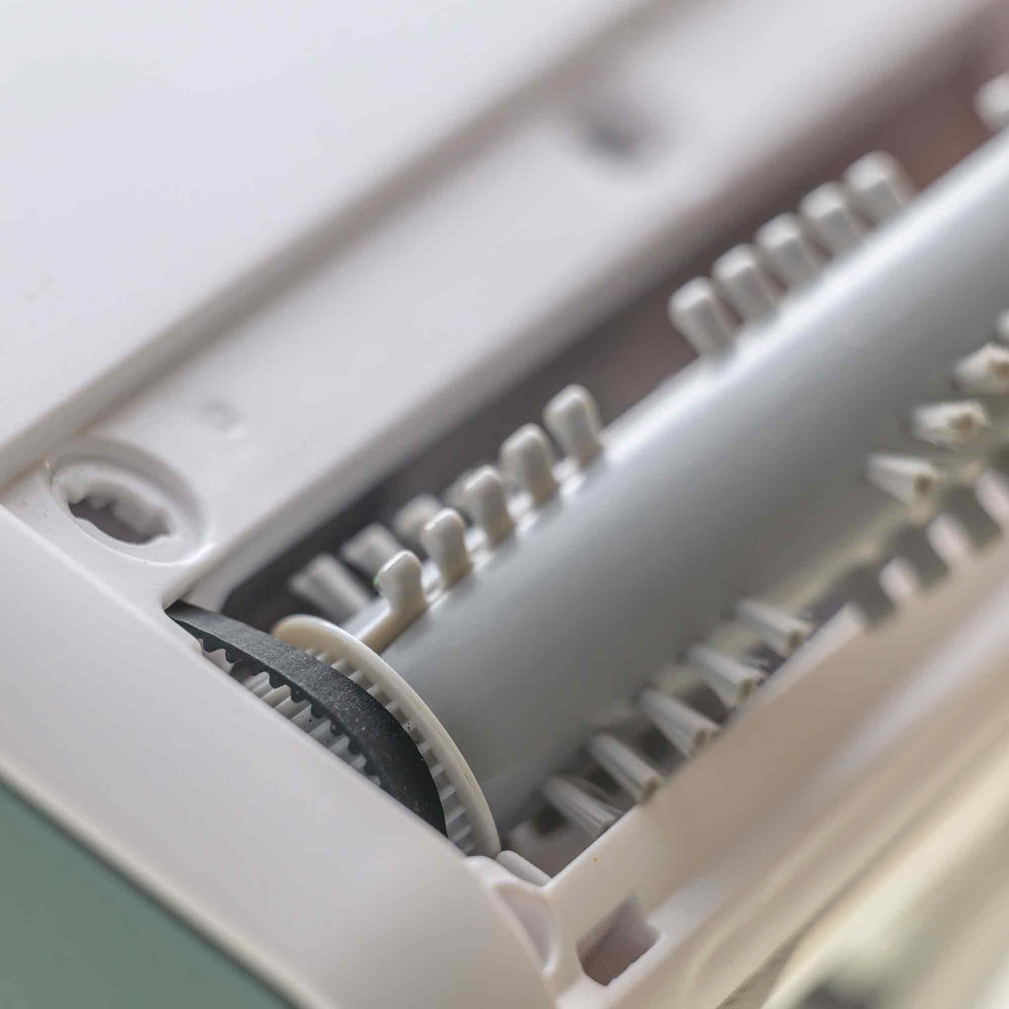 Zbliżenie na wewnętrzne części białego urządzenia czyszczącego. Widoczny jest czarny pasek, koła zębate i szczotka rolkowa.