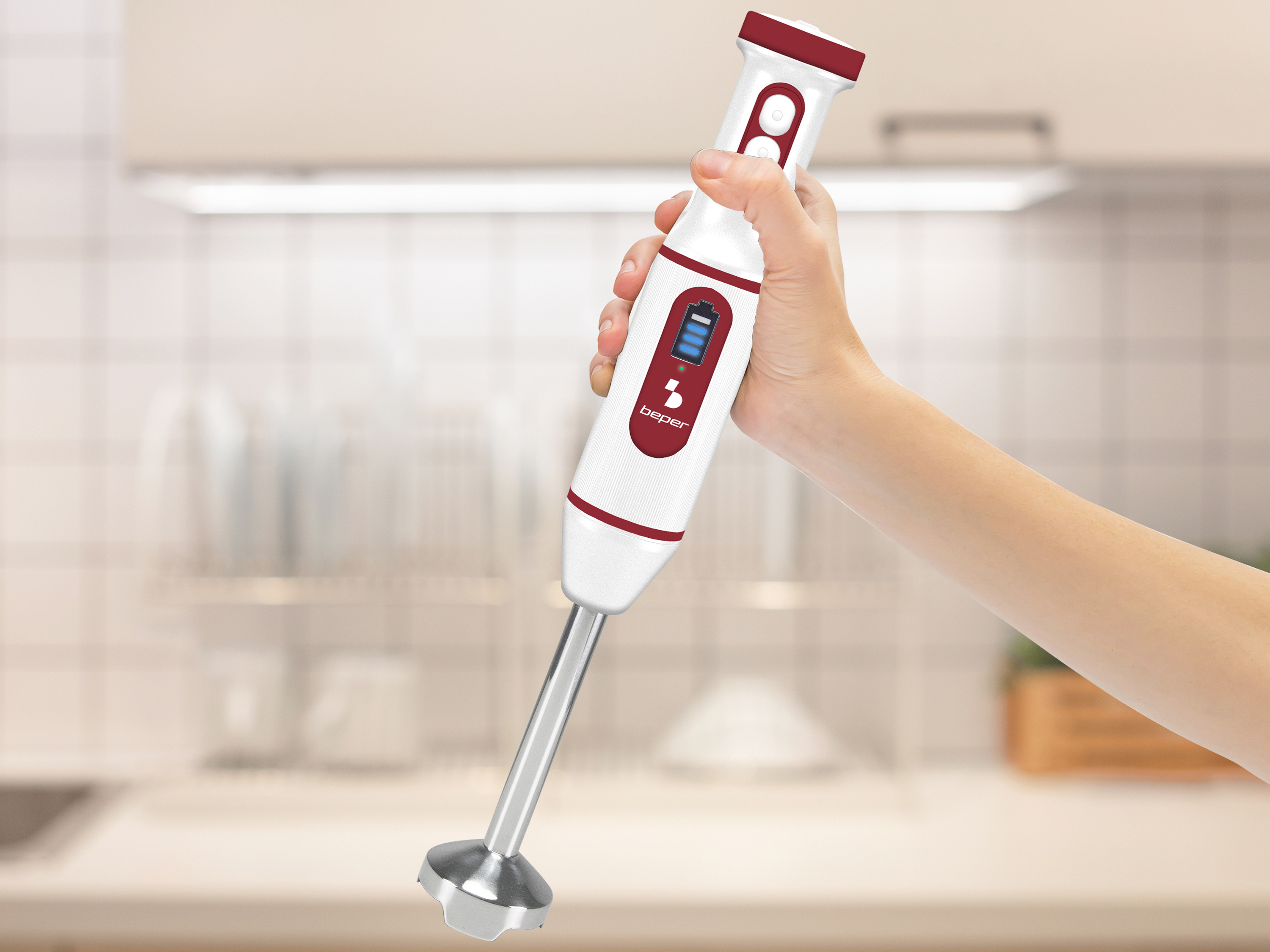 Una mano tiene un frullatore a immersione bianco e rosso. Sfondo: cucina con bancone e armadi sfocati.