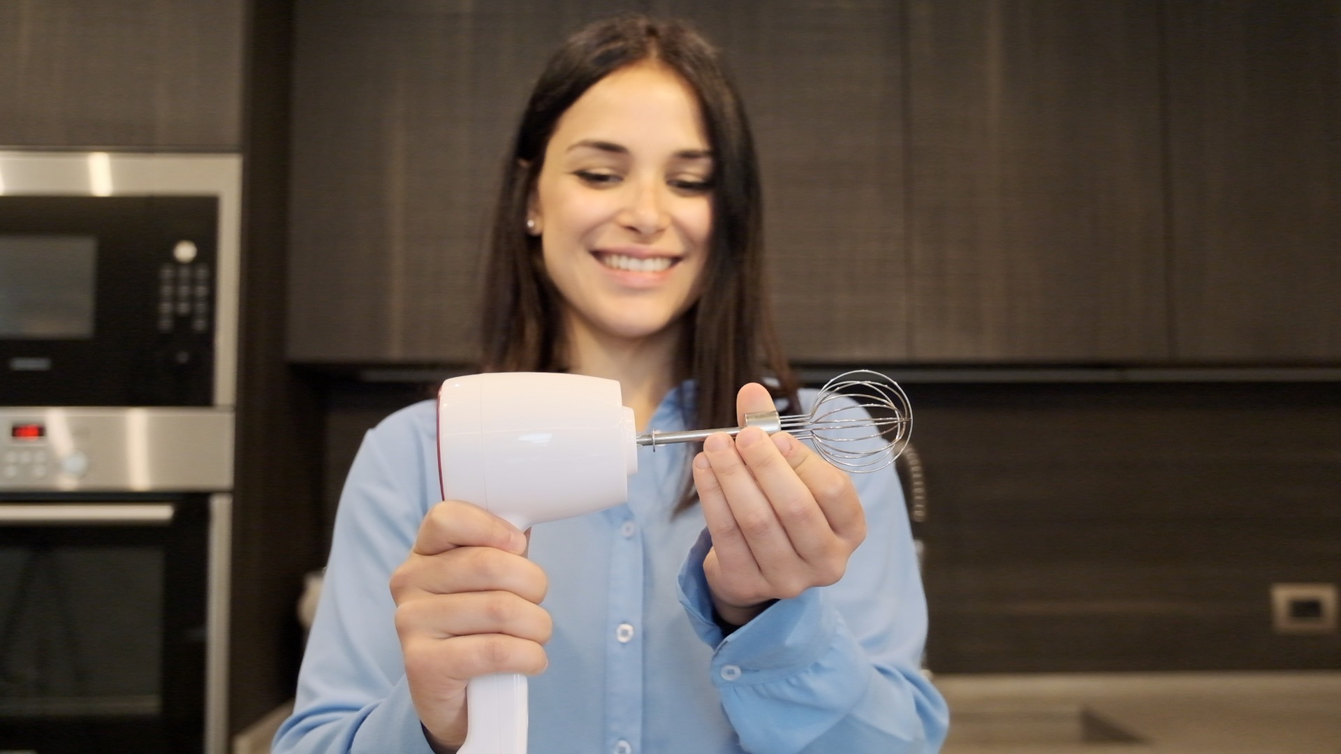 Donna sorride, tiene in mano un frullatore a immersione bianco e una frusta. Sfondo cucina con microonde e mobili scuri.