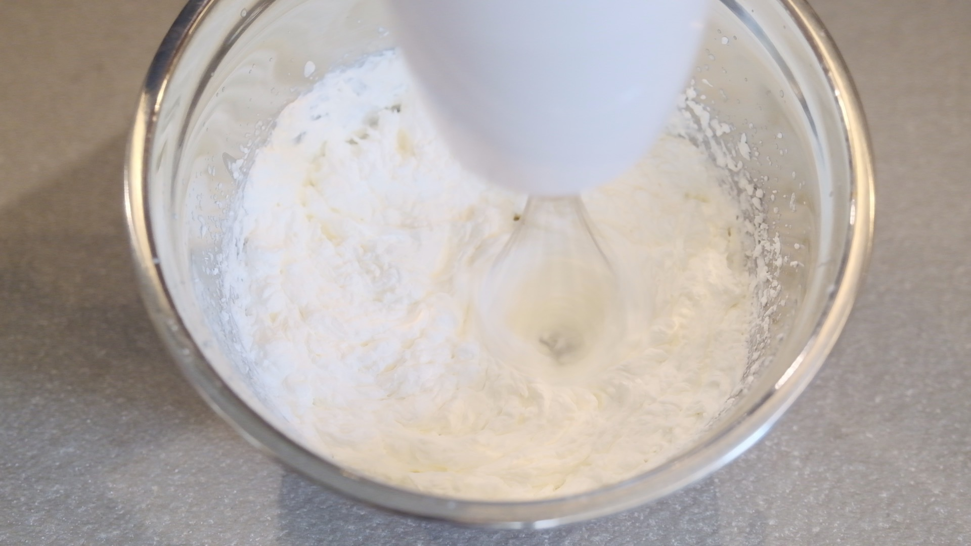Un mixer a mano bianco sta mescolando la panna in una ciotola di vetro trasparente. La panna è soffice e bianca. Lo sfondo è una superficie grigia.