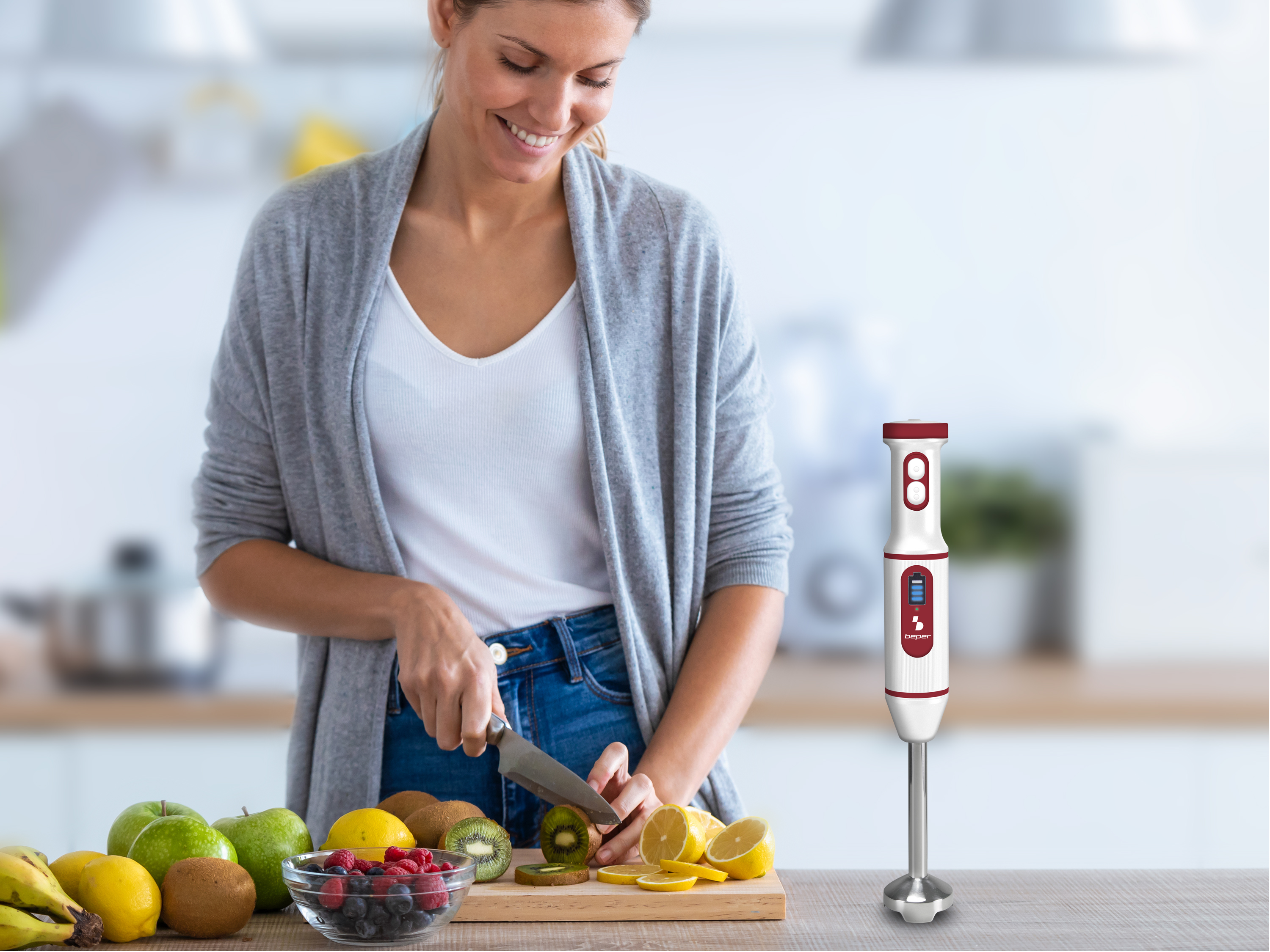 Donna taglia kiwi con coltello su tagliere, circondata da frutta e frullatore.