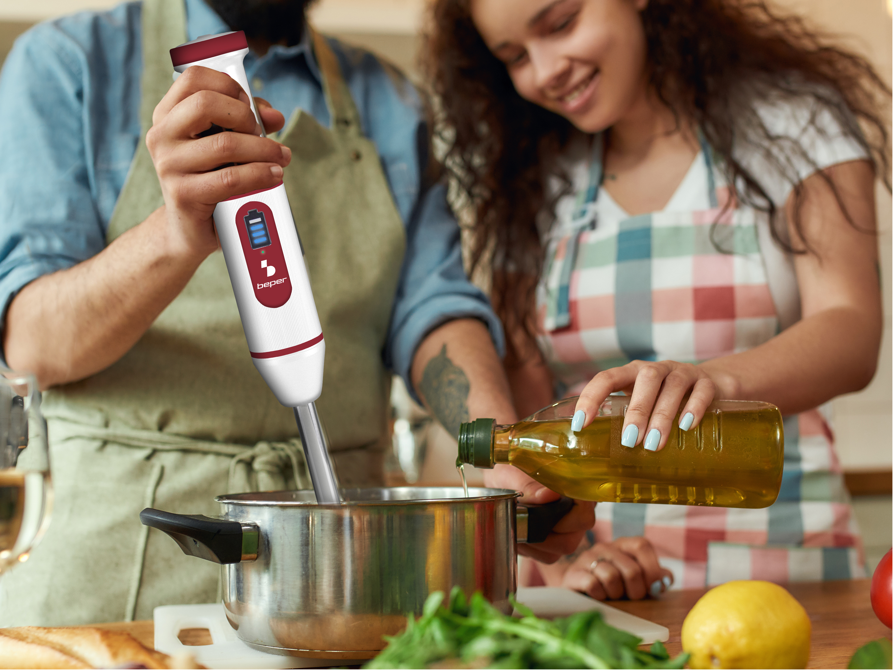 Una persona usa un frullatore in una pentola, mentre un'altra versa olio. Limone e basilico sul tavolo.