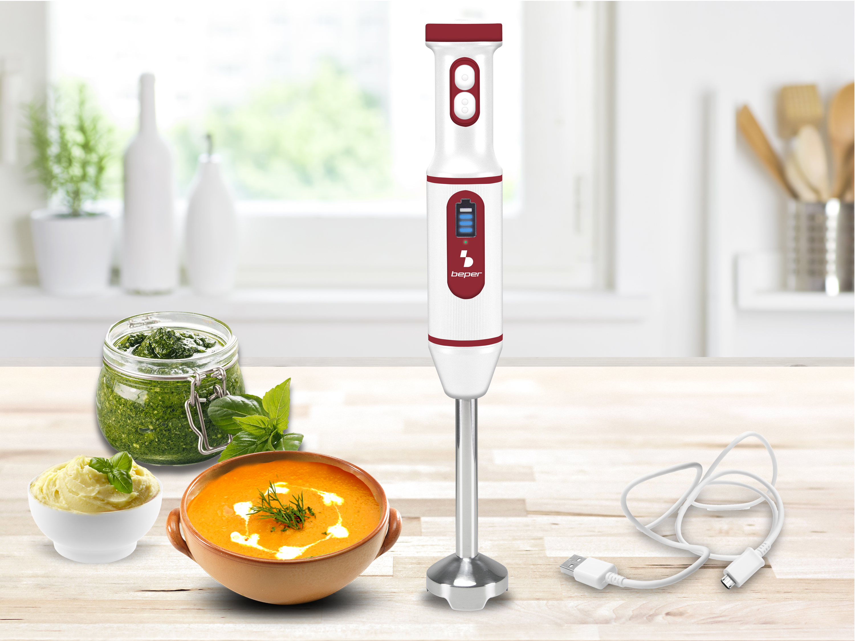 Un frullatore a immersione bianco e rosso è su un tavolo di legno con zuppa, pesto e un cavo di ricarica.