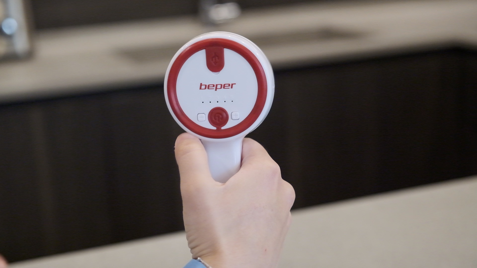 Una mano tiene in mano un dispositivo bianco e rosso con la scritta 'beper'. Lo sfondo ha un piano di lavoro scuro.