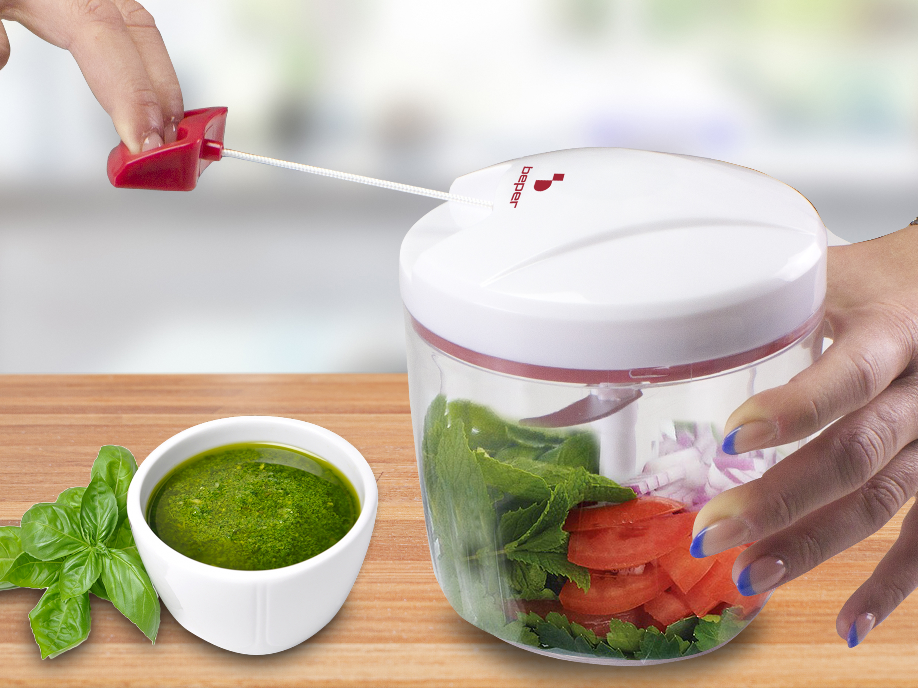 Una mano tira un cavo rosso attaccato a un tritatutto bianco. Dentro, verdure. Una ciotola di pesto è di lato.