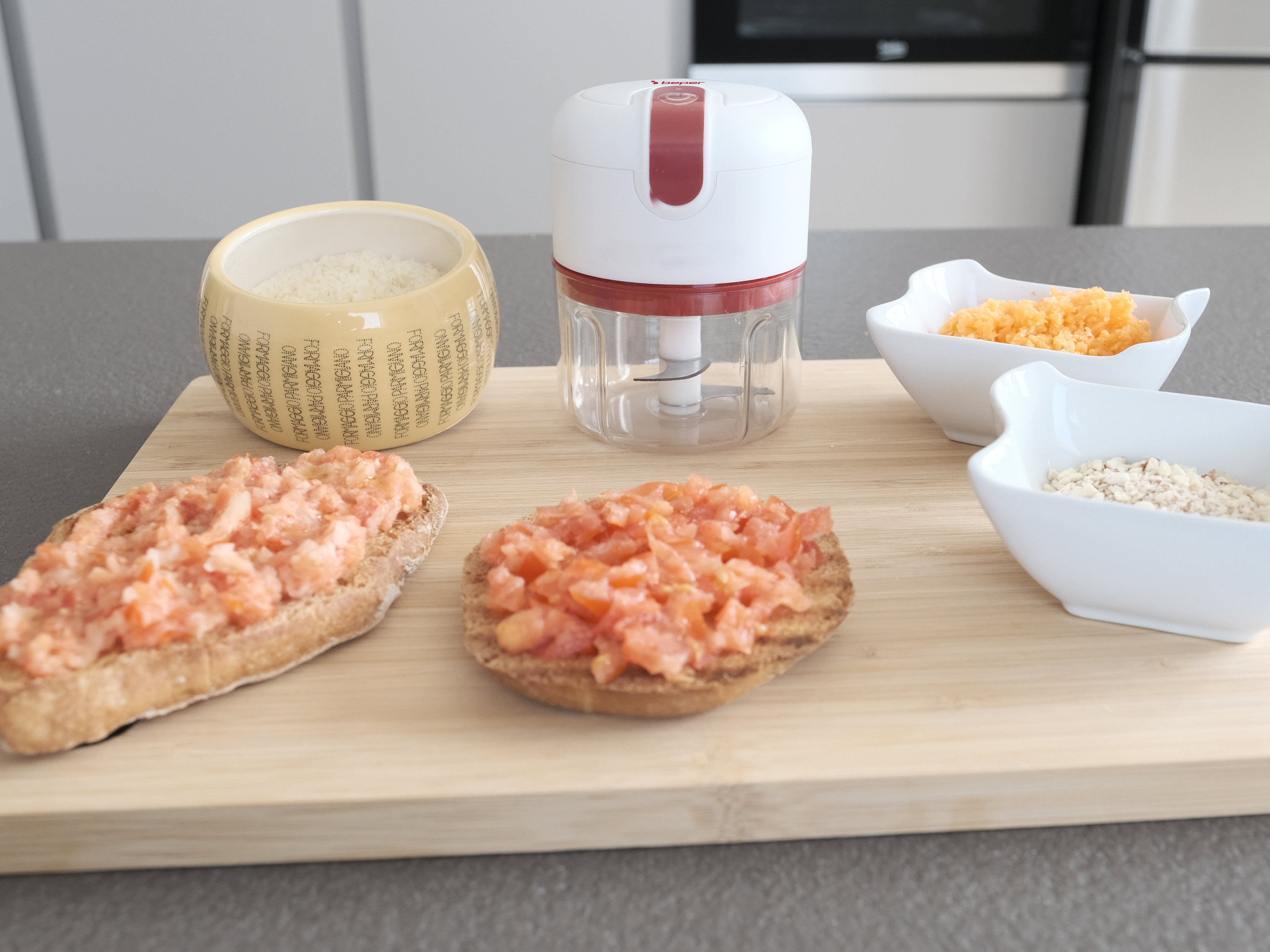 Un tagliere con bruschetta al pomodoro, ciotole di formaggio grattugiato e ingredienti e un robot da cucina.