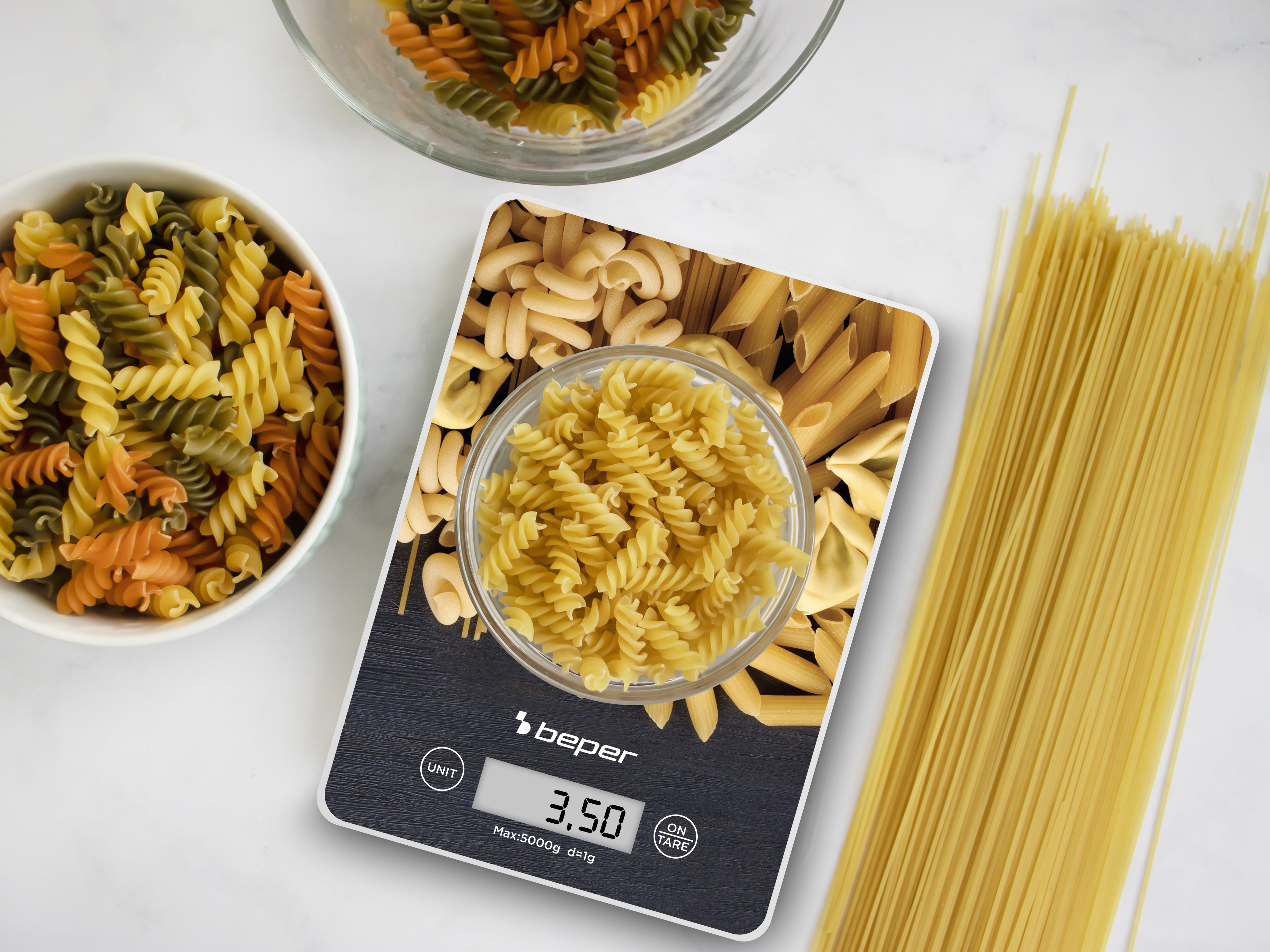 Pasta su una bilancia, con ciotole di pasta e spaghetti. La bilancia indica 3,50. Sono visibili varie forme e colori di pasta.