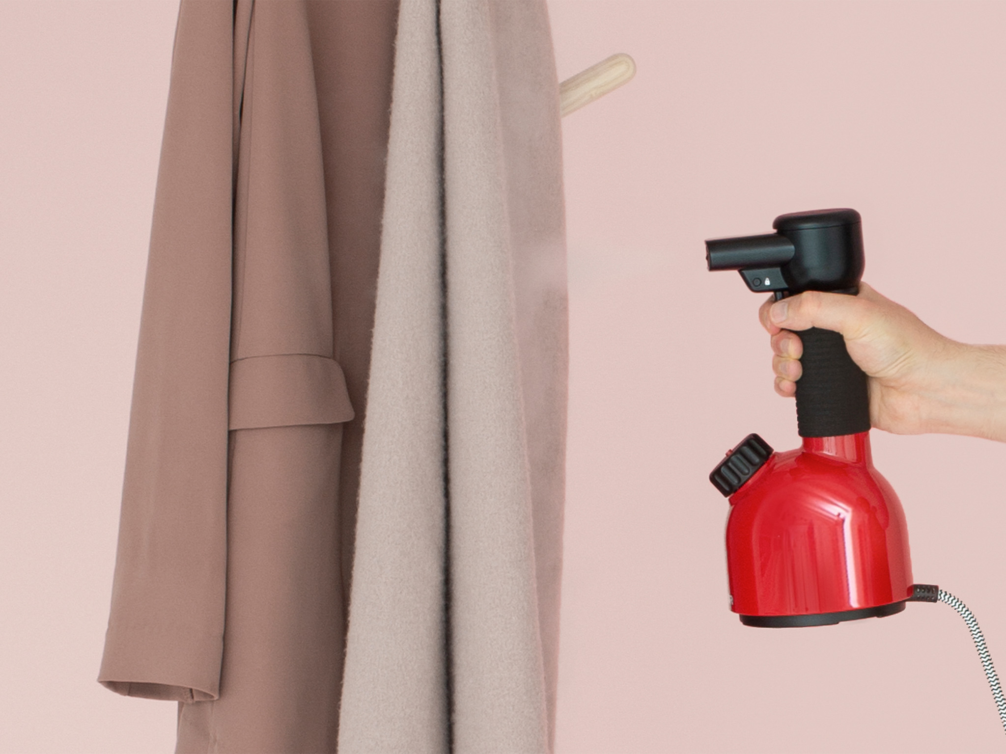 Eine Person dampft einen Mantel mit einem roten Dampfgarer. Mäntel hängen an einem Holzbügel vor rosa Hintergrund.