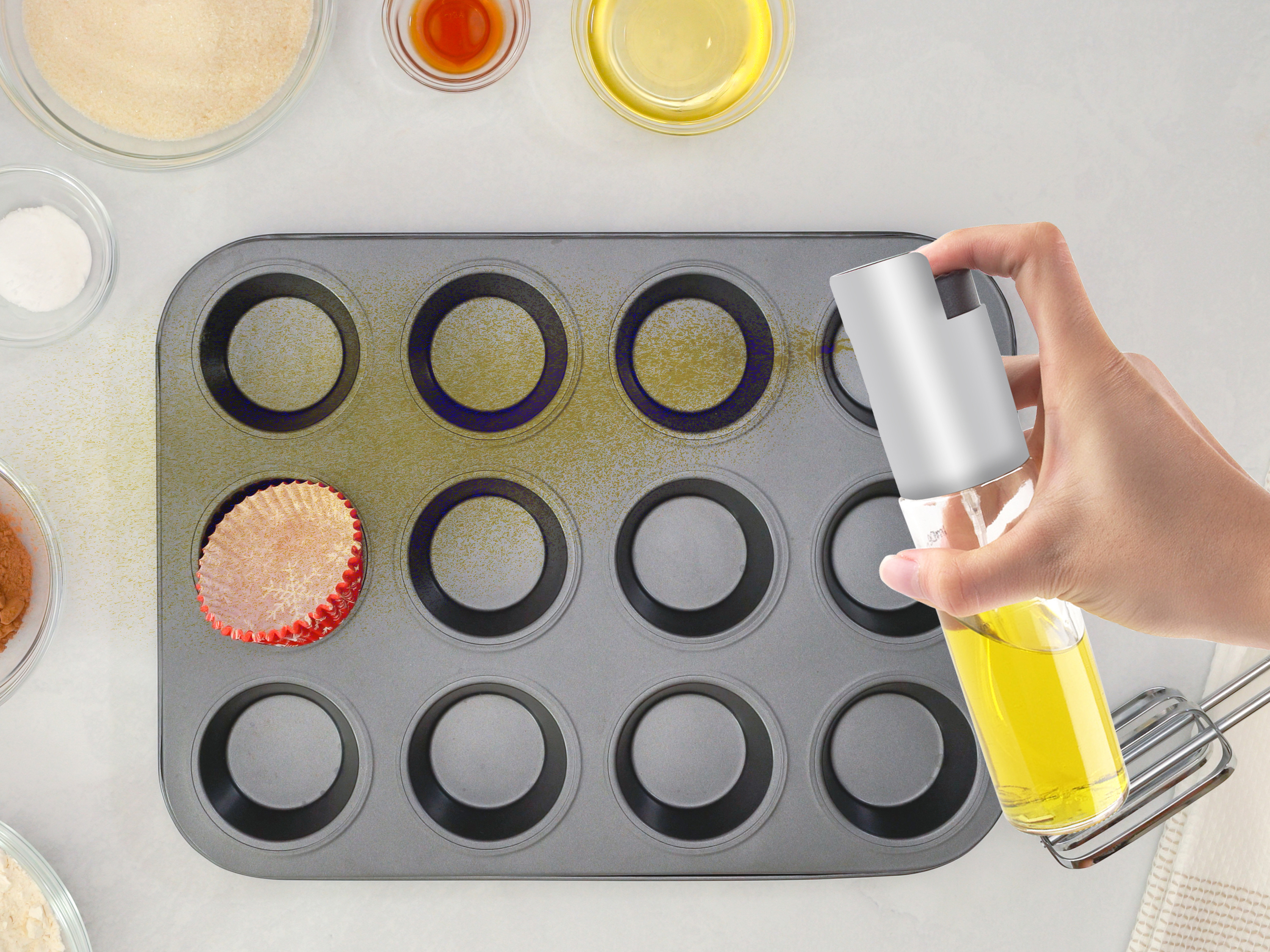 Una mano spruzza olio in una teglia per muffin. Ingredienti e frusta sono sulla superficie bianca.