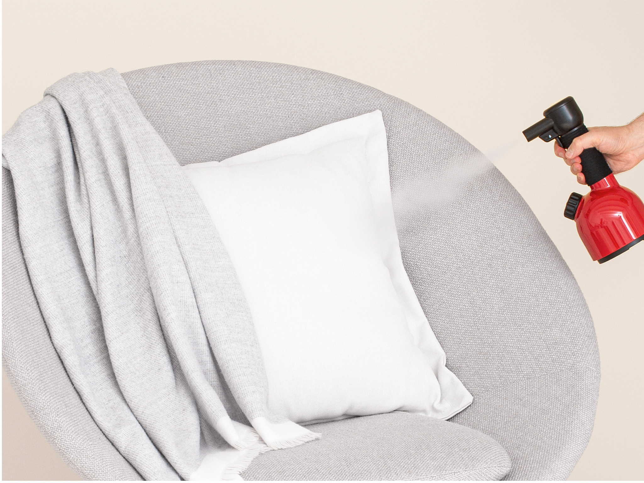Un fauteuil gris avec un oreiller et une couverture blancs. Une main vaporise le fauteuil avec un vaporisateur rouge et noir.
