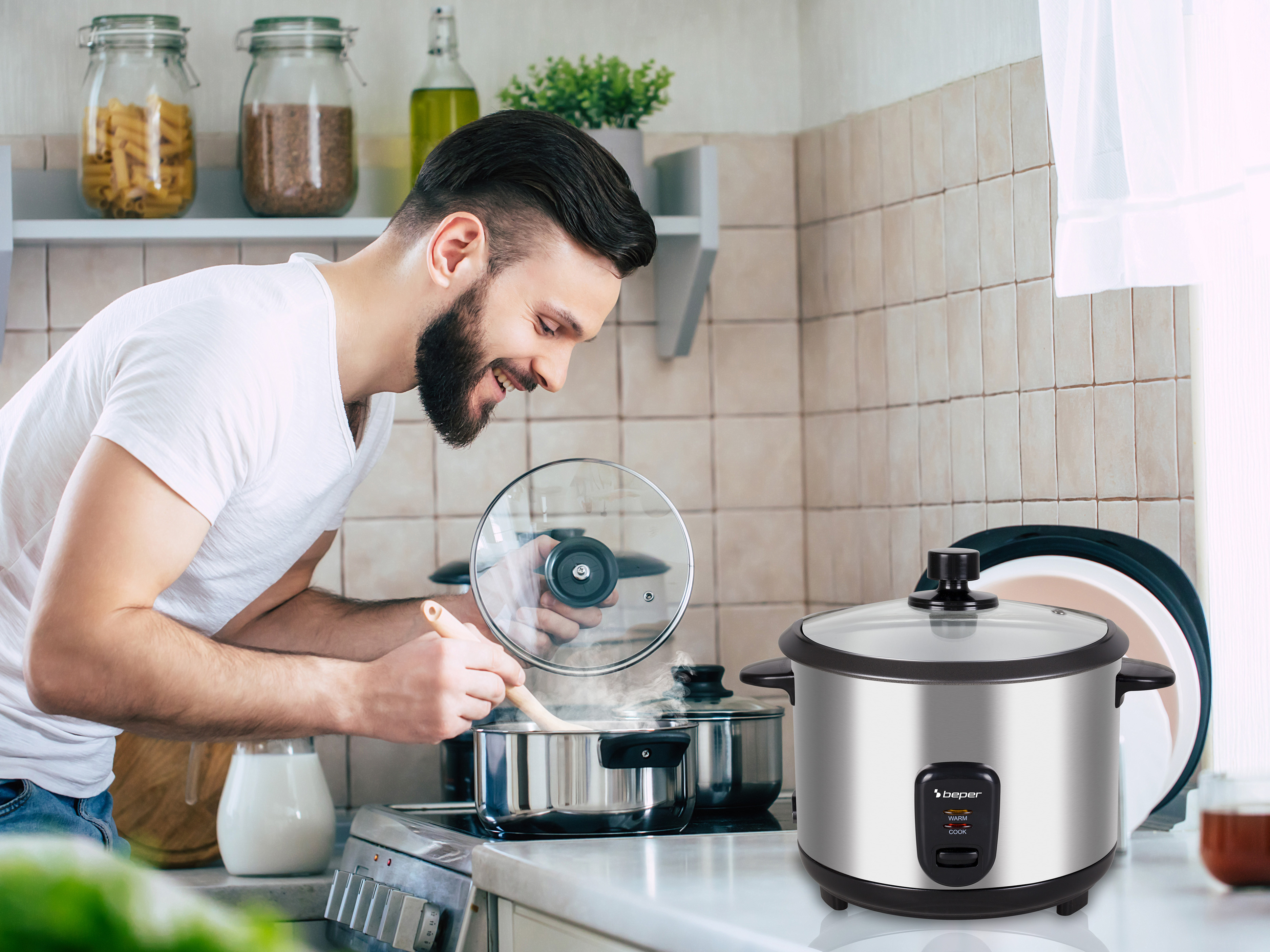 Uomo che cucina, vapore che sale da una pentola. Cuociriso in primo piano. Ambiente cucina con barattoli e piastrelle.