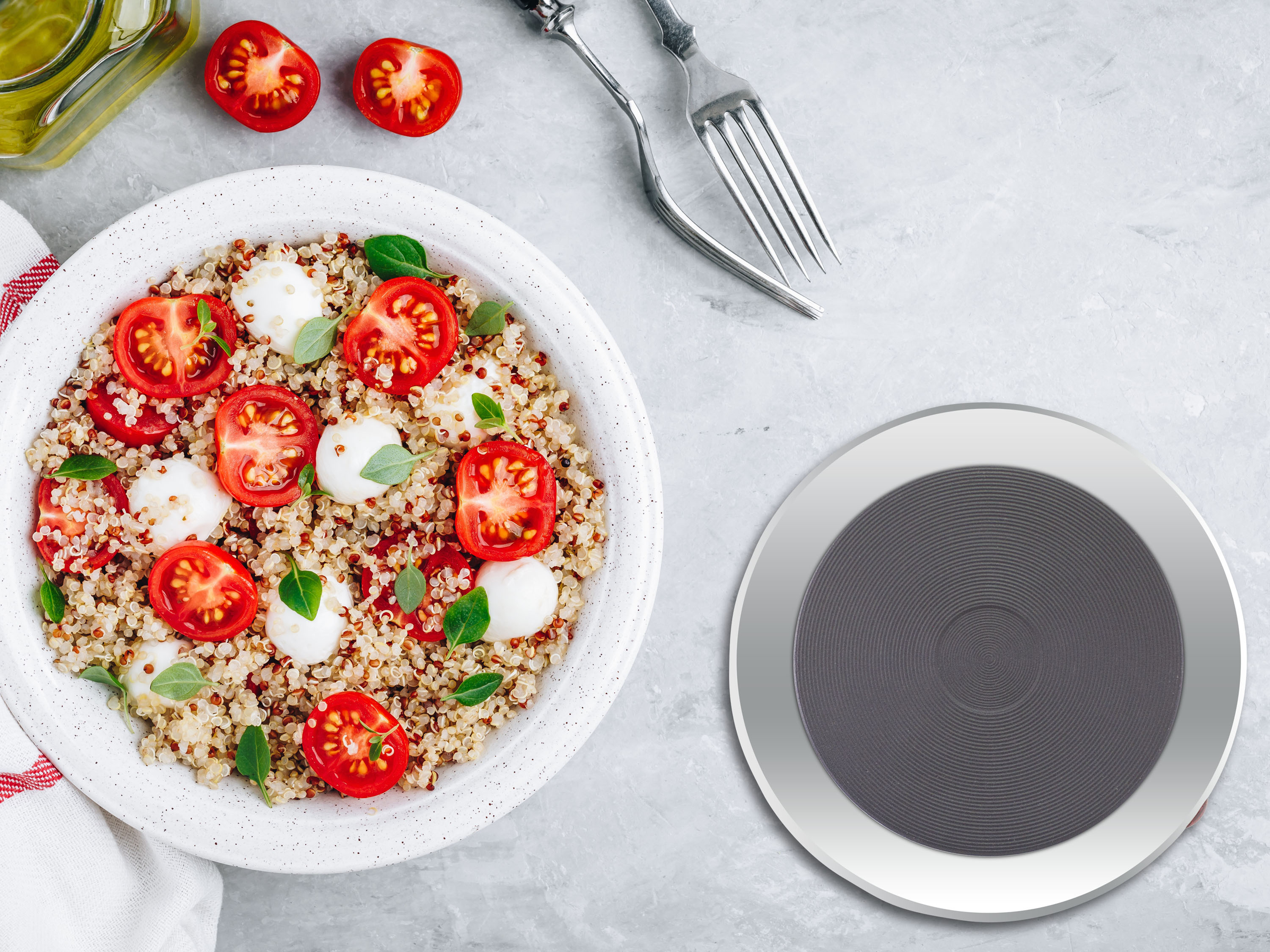 Un'insalata di quinoa con pomodori e mozzarella in una ciotola bianca. Forchette, olio e sottobicchiere.