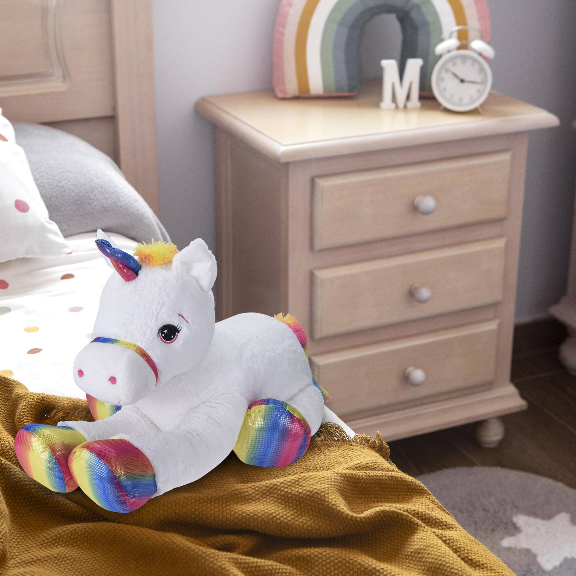 Un peluche de unicornio blanco con acentos de arcoíris en una cama con una mesita de noche en el fondo.