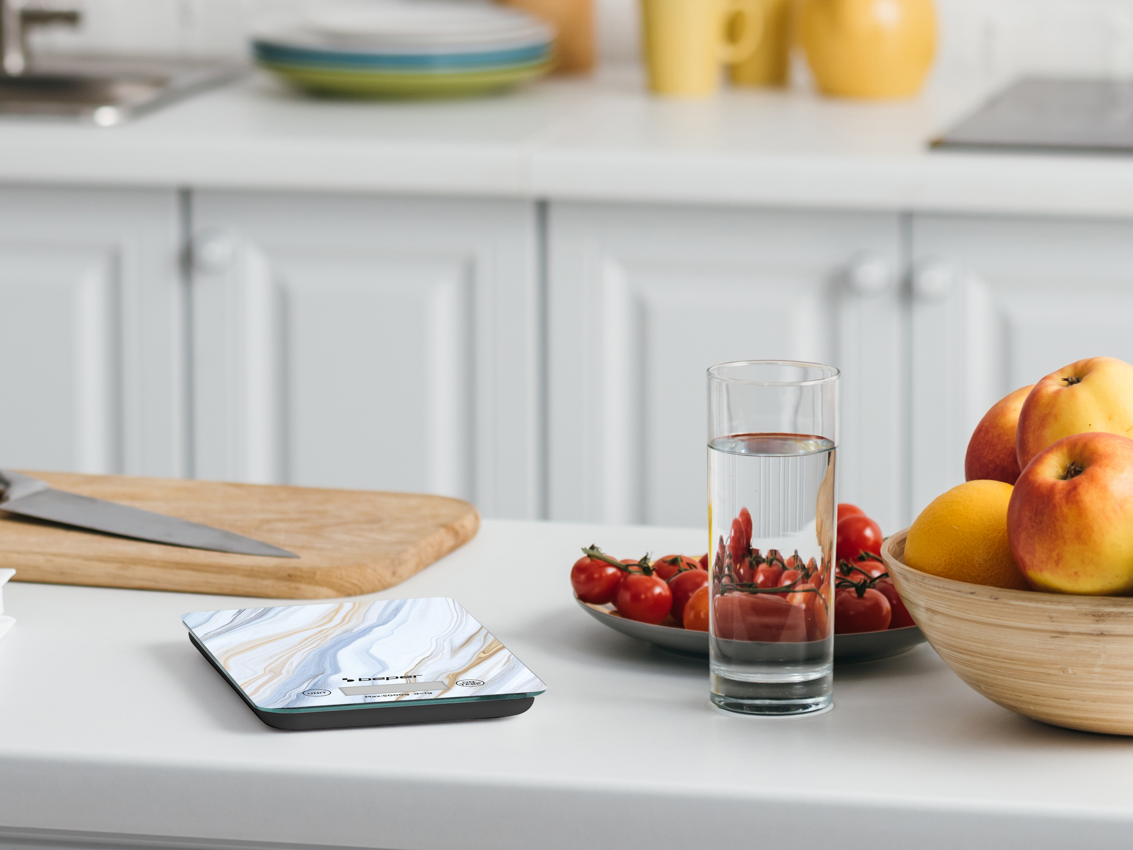 Scena di cucina. Bilancia, pomodori, mele, limone, bicchiere d'acqua, coltello, tagliere, piatti e armadi bianchi.