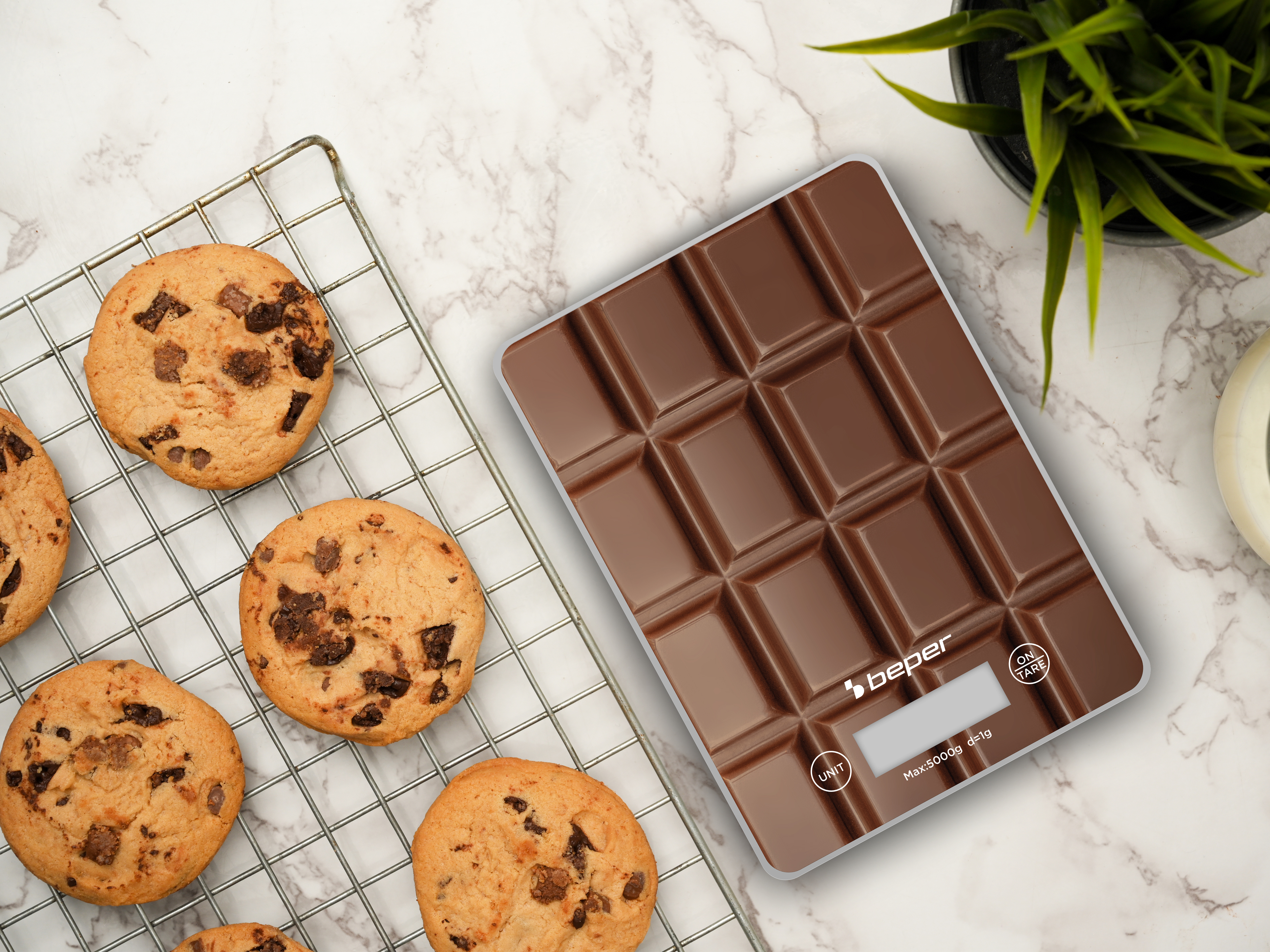 Biscotti su griglia, bilancia al cioccolato e pianta su sfondo marmoreo.