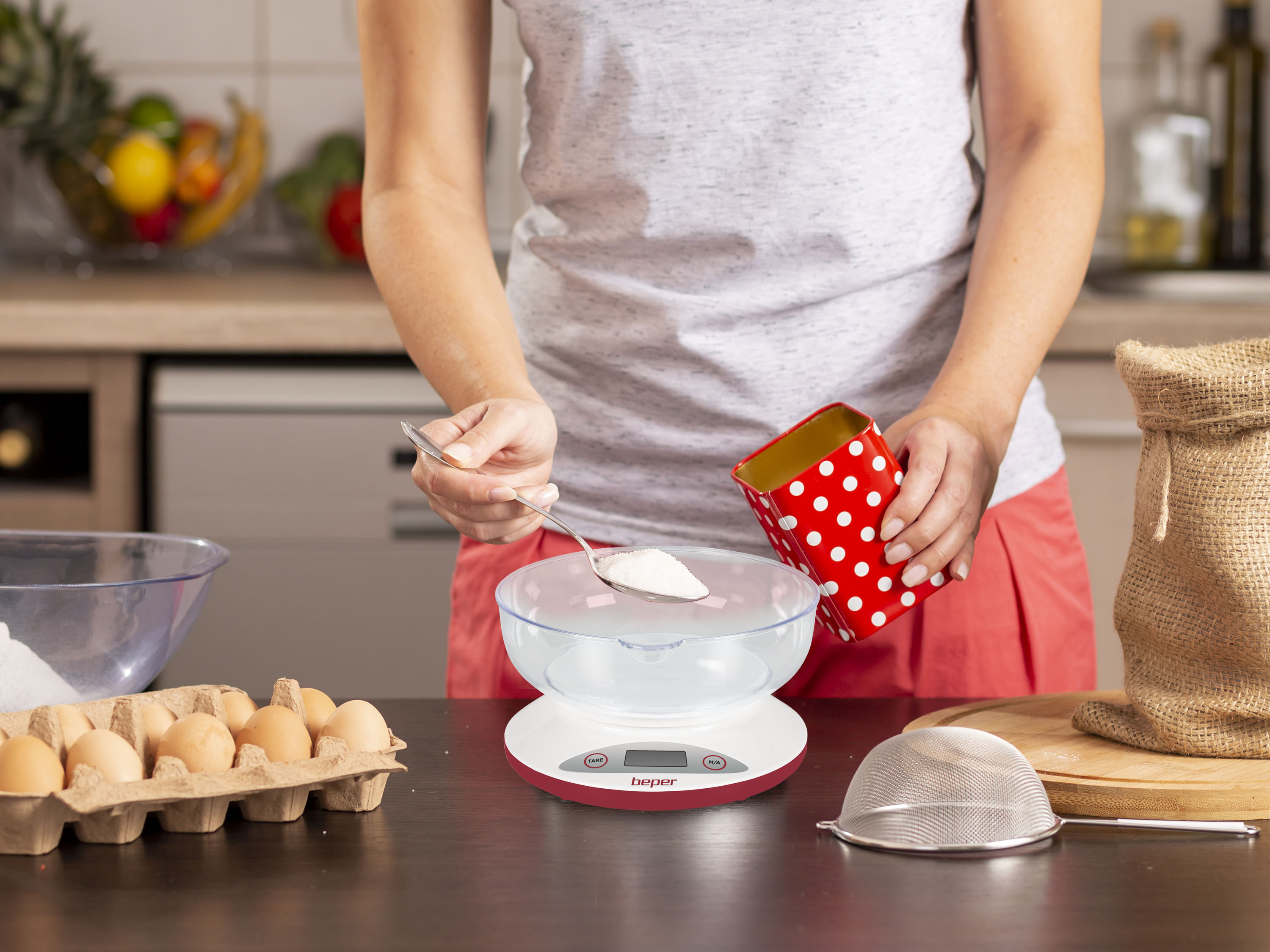 Una persona sta misurando la farina con un cucchiaio in una ciotola su una bilancia da cucina. Altri ingredienti sono visibili.