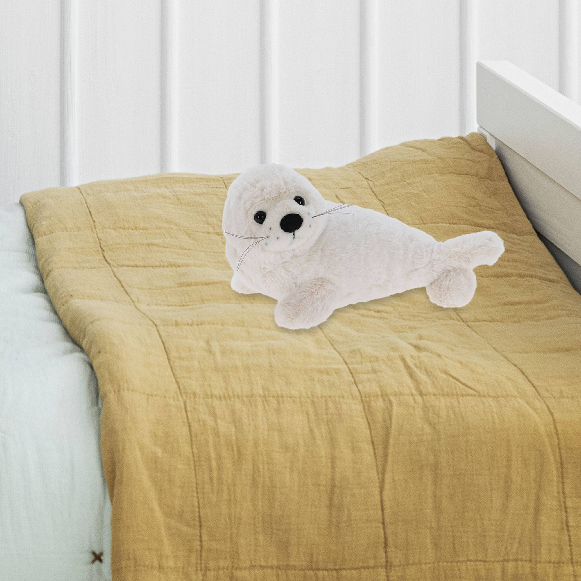 Un peluche de foca blanca yace sobre una manta amarilla, colocado en una cama. Pared blanca al fondo.