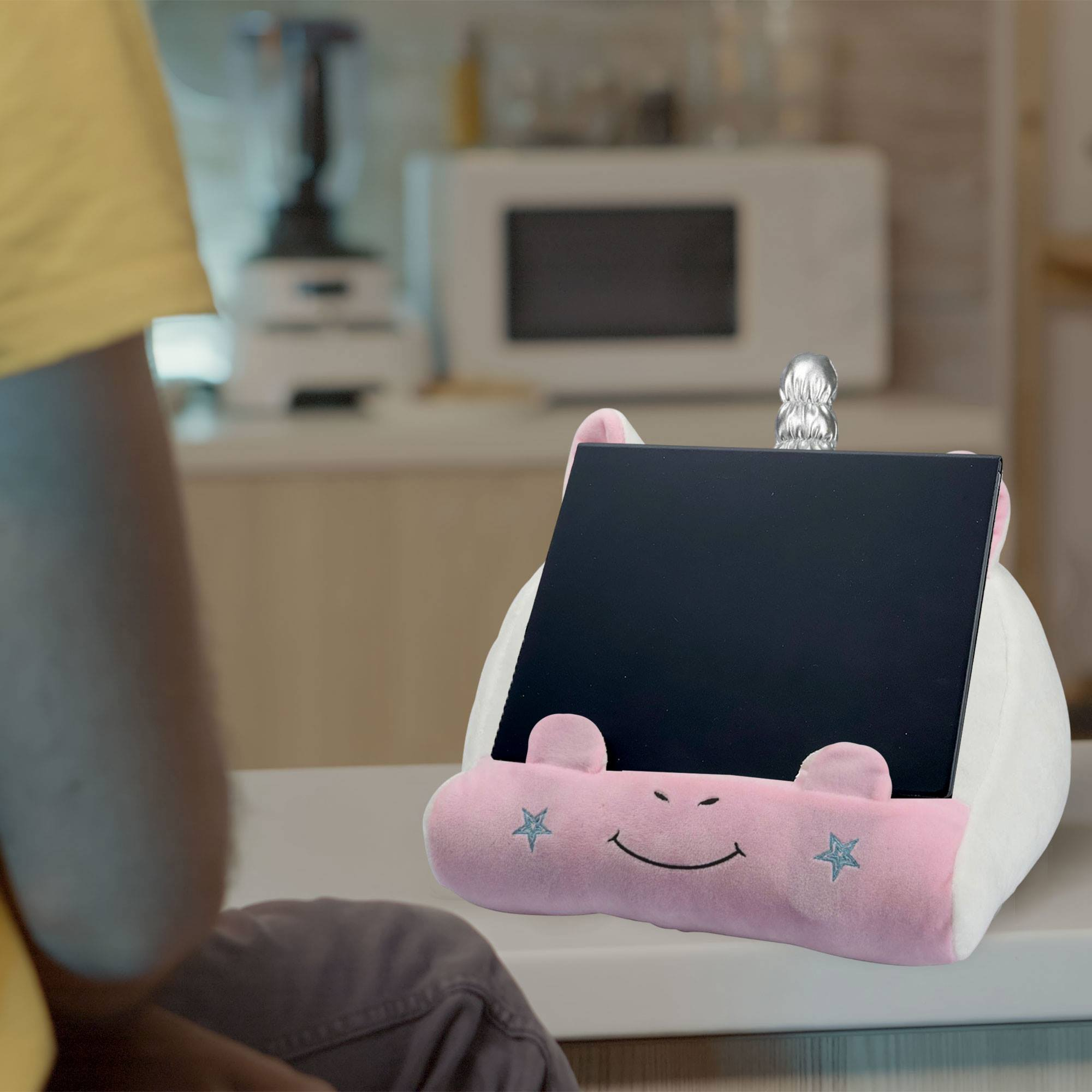 Una tableta en un soporte con forma de unicornio está sobre una superficie blanca. Una persona está sentada delante. Un microondas está detrás.