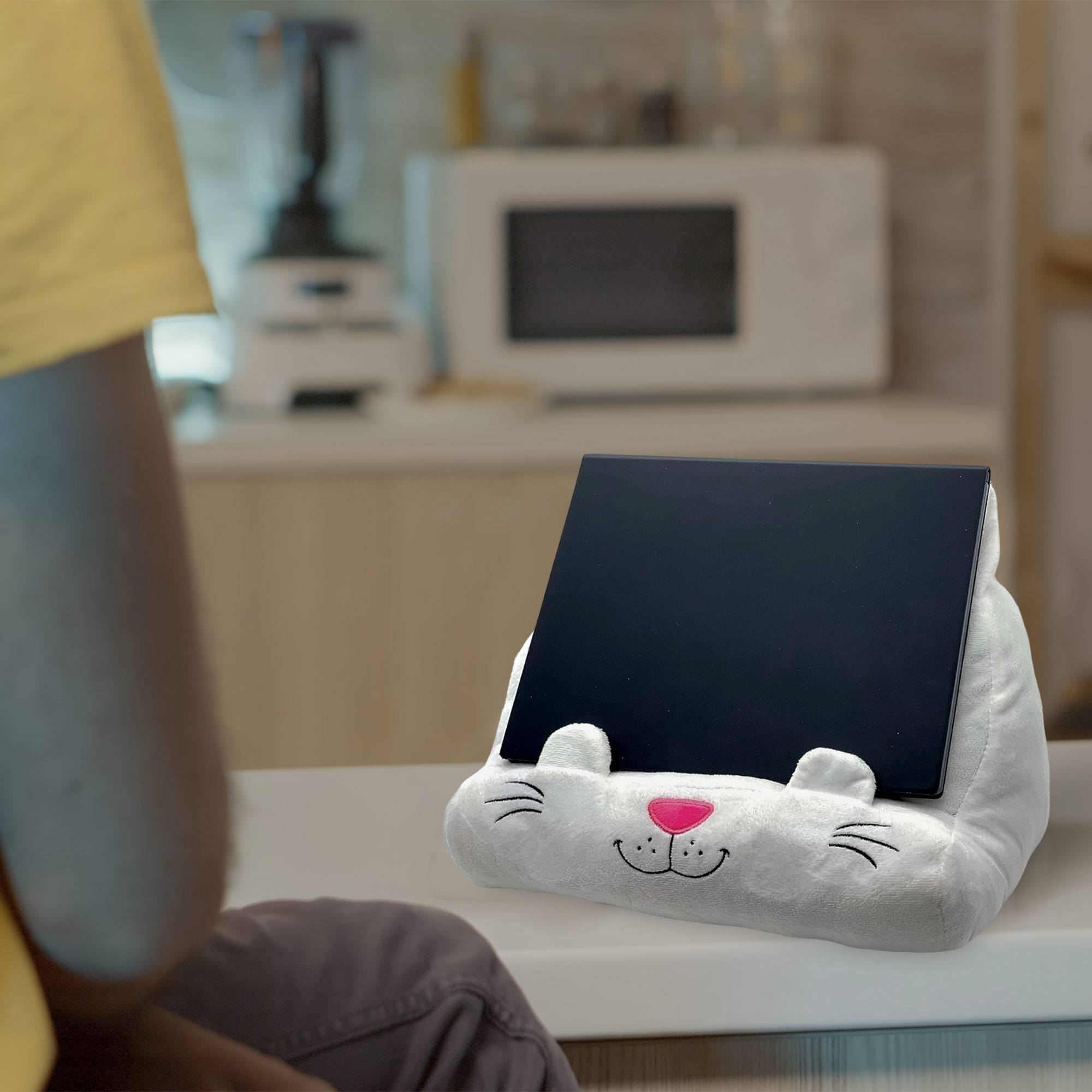 Una tableta descansa en un soporte blanco con forma de gato. Una persona es visible en primer plano. Un microondas está al fondo.