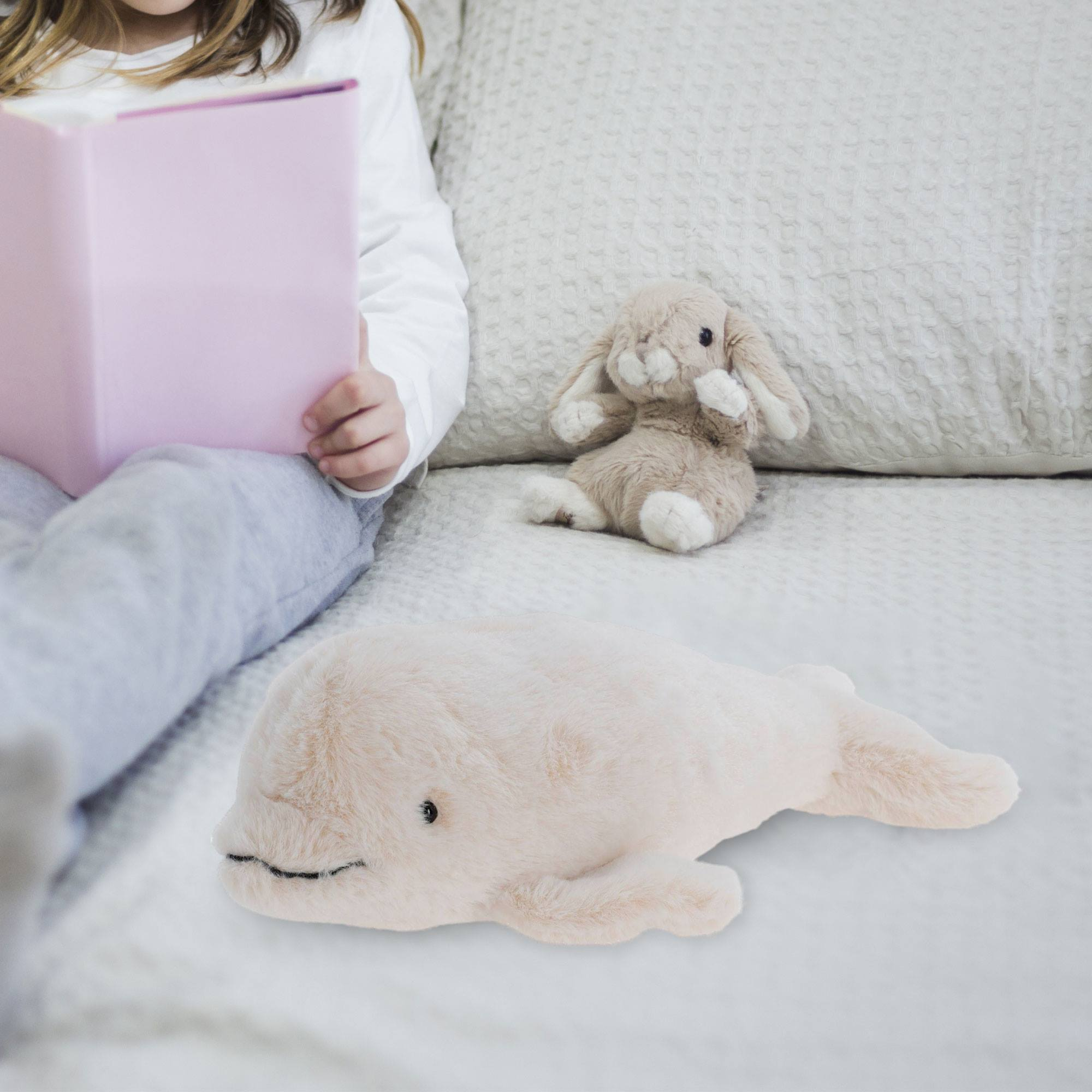 Un niño lee un libro rosa en una cama. Un conejito de peluche y un juguete de ballena beige están en la cama.