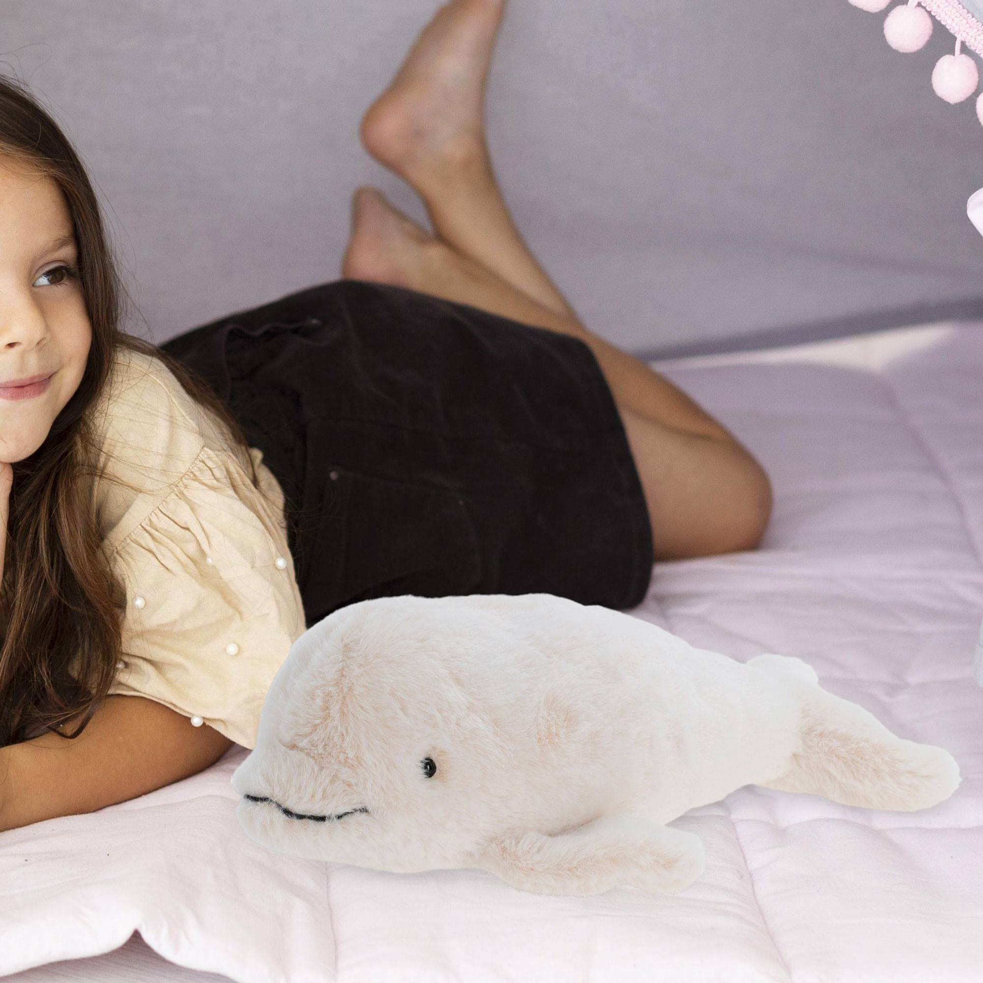 Una niña está acostada sobre una manta rosa, un peluche de ballena blanca frente a ella. Usa una camisa beige.