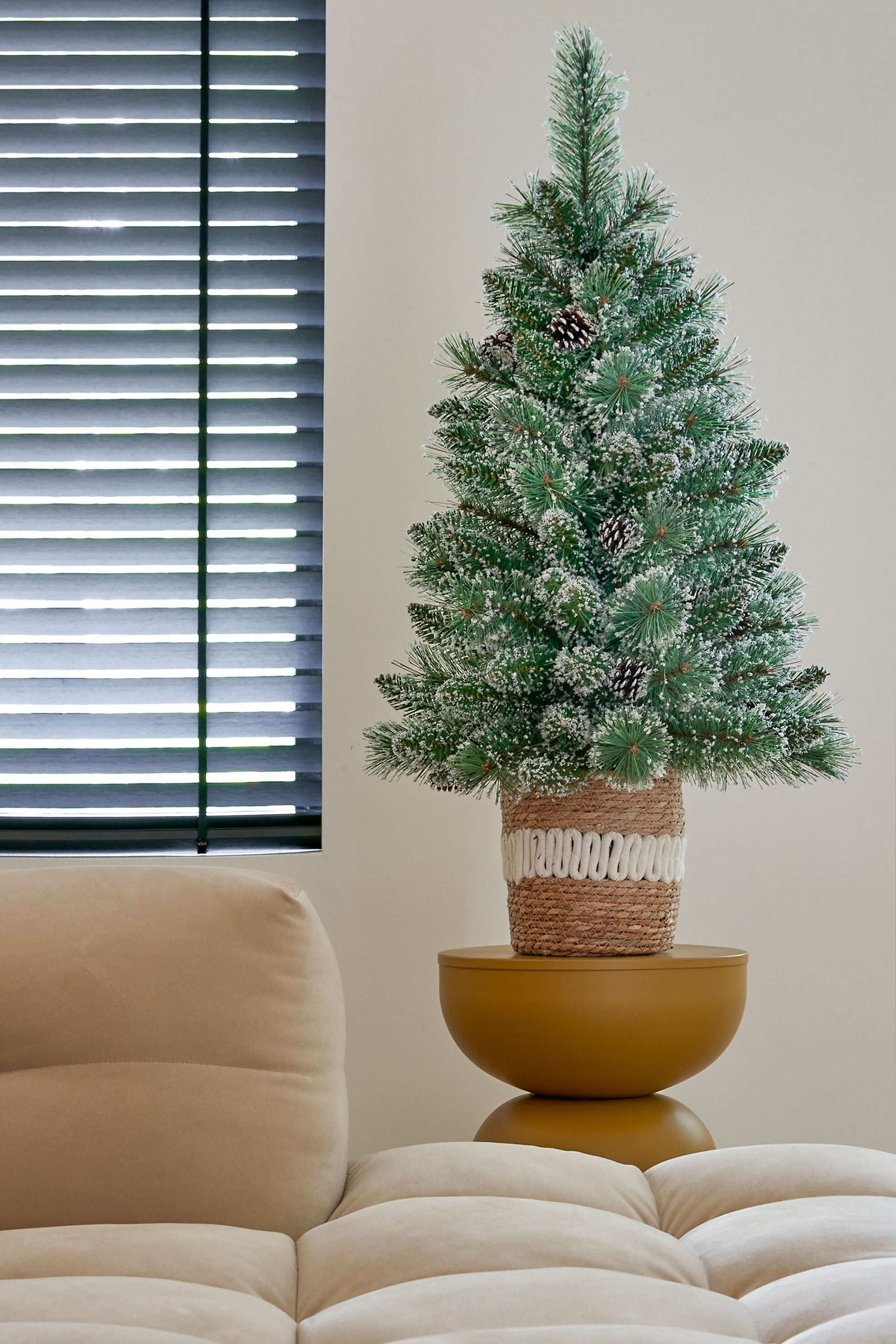 Un petit sapin de Noël avec effet neige, sur une table jaune. Stores et un canapé beige en arrière-plan.