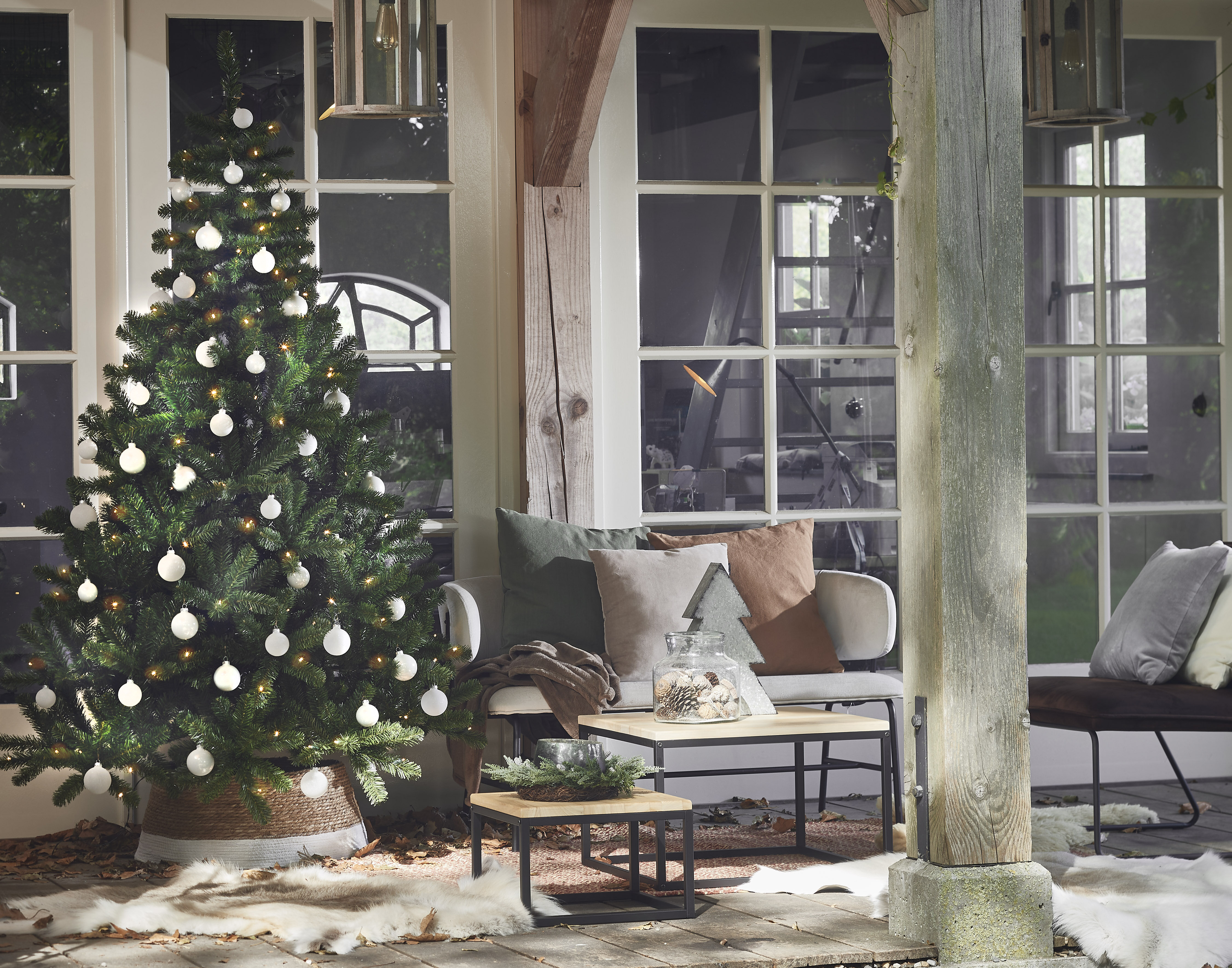 Sapin de Noël avec ornements blancs, canapé, coussins et tables en bois sur un porche.