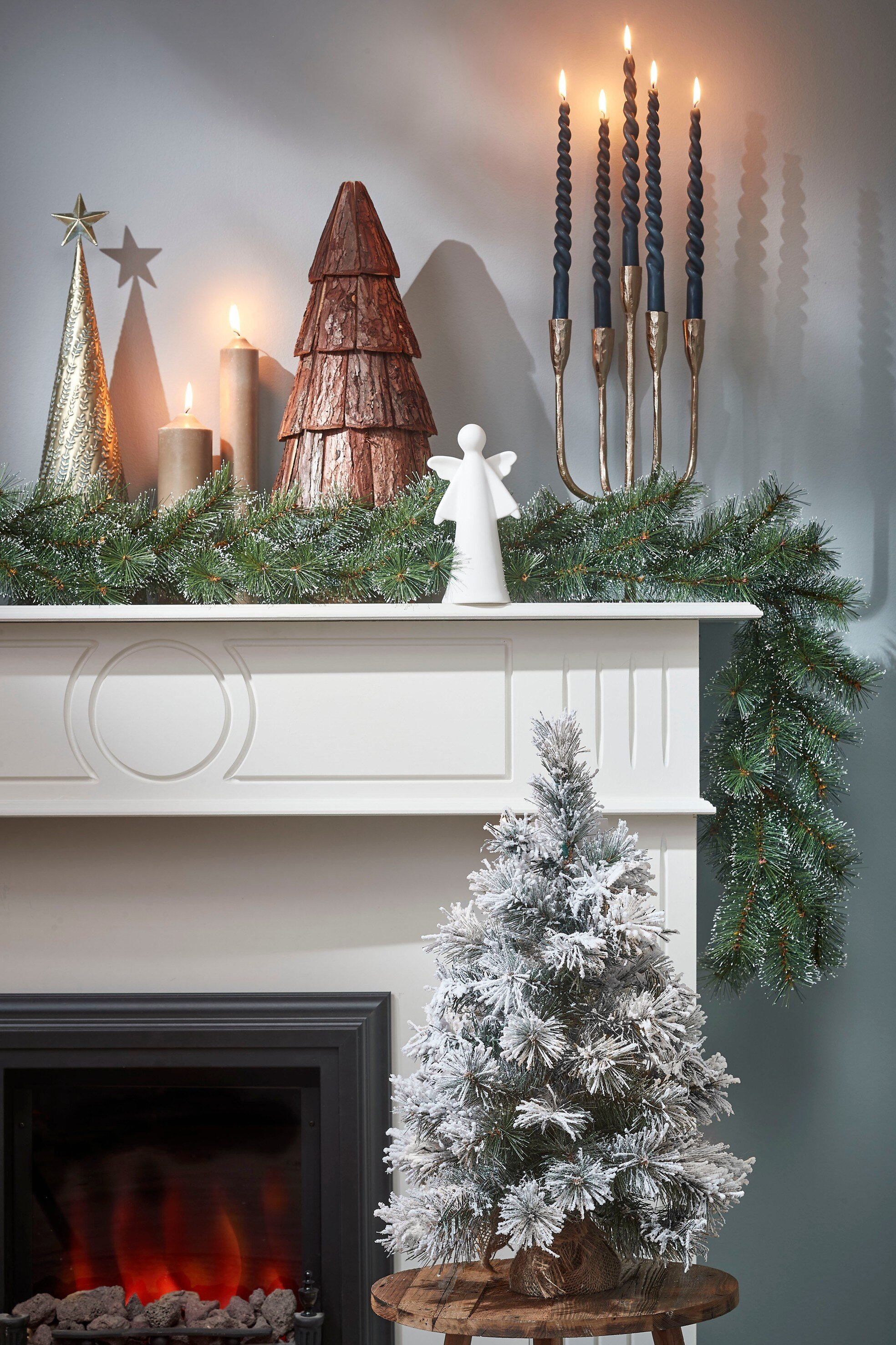 Un manteau décoré avec des bougies, un ange et une guirlande. Un arbre givré est devant une cheminée.