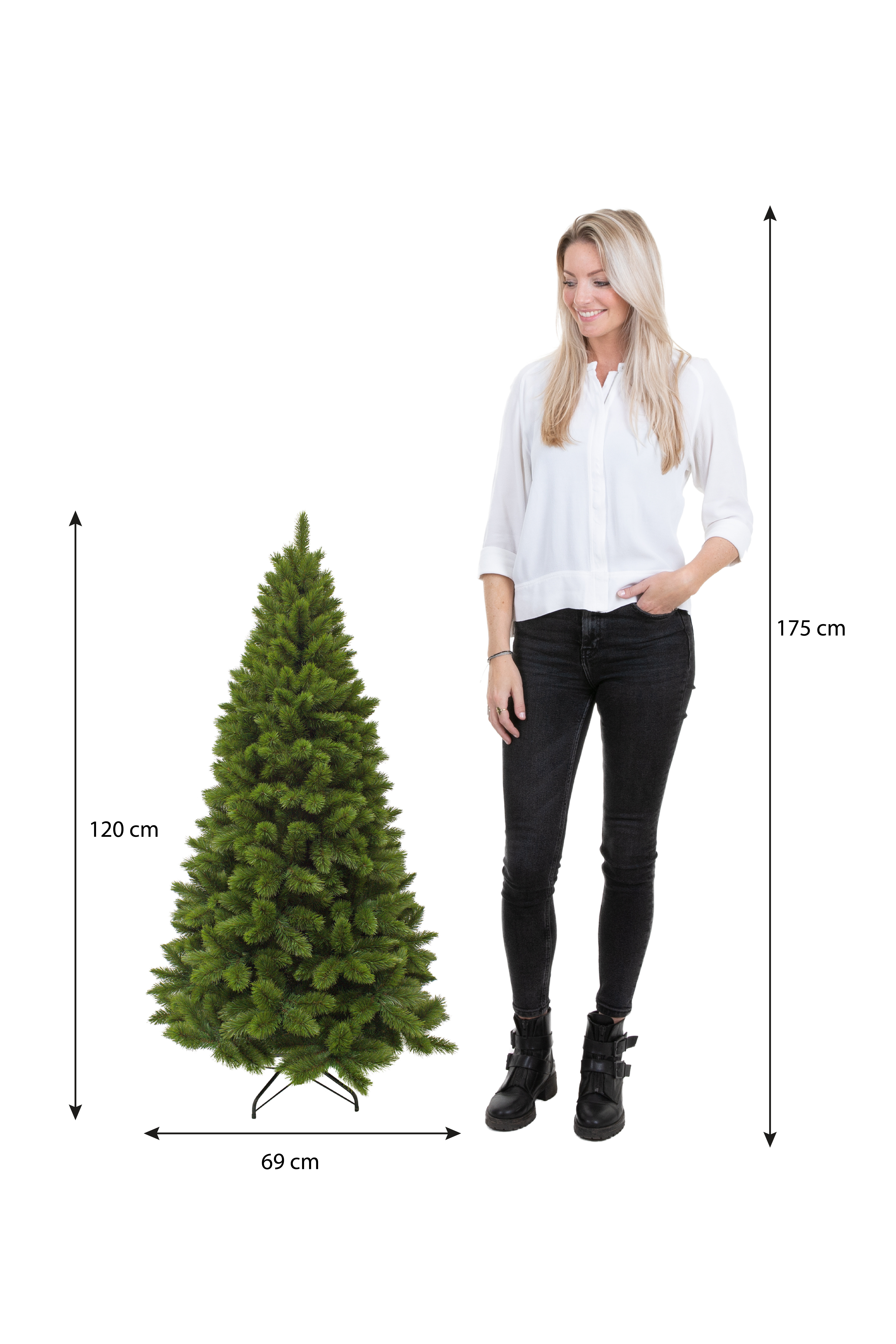 Une femme se tient à côté d'un sapin de Noël. L'arbre est vert. La femme porte un jean noir et une chemise blanche.