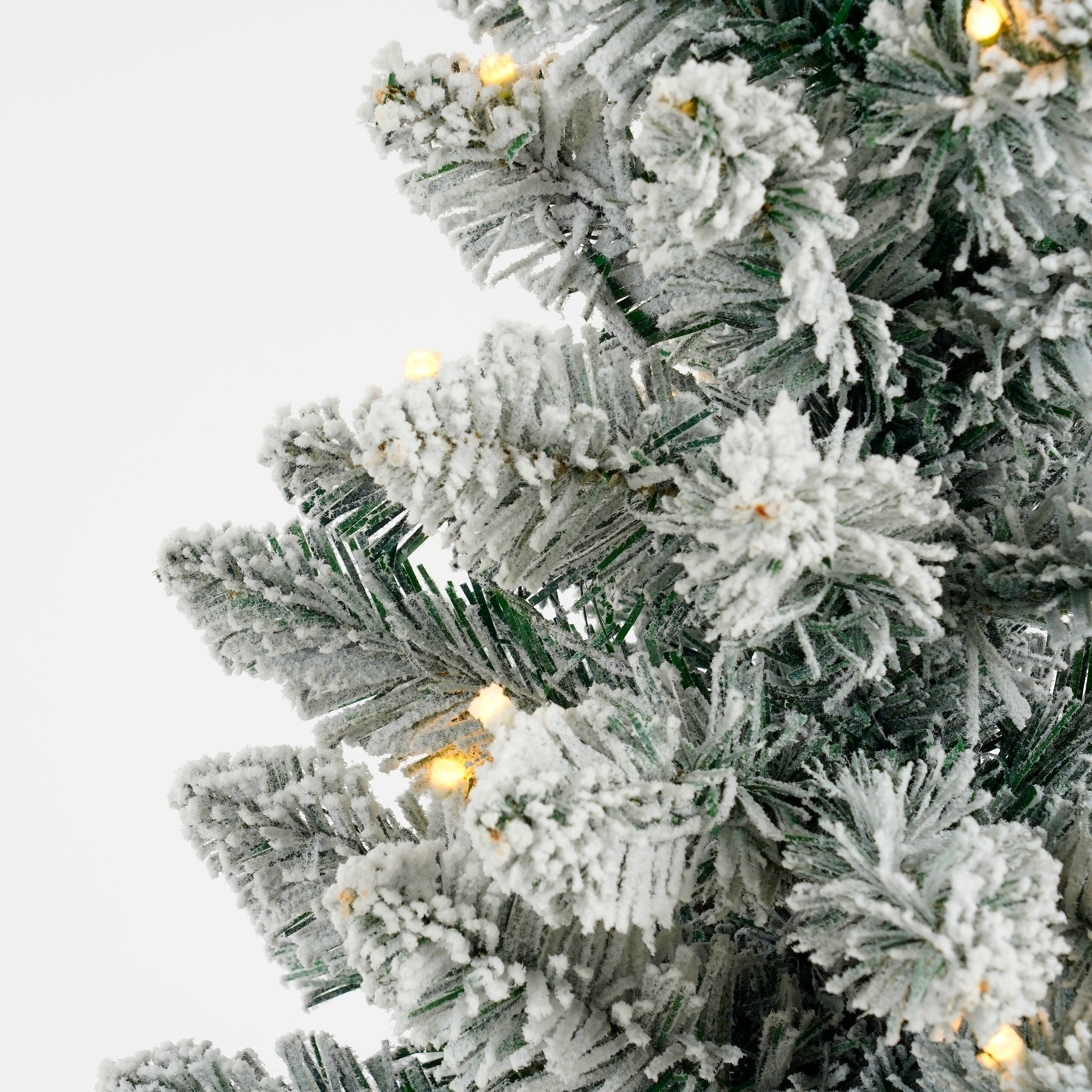 Gros plan d'un sapin de Noël enneigé avec de petites lumières jaunes sur fond blanc.