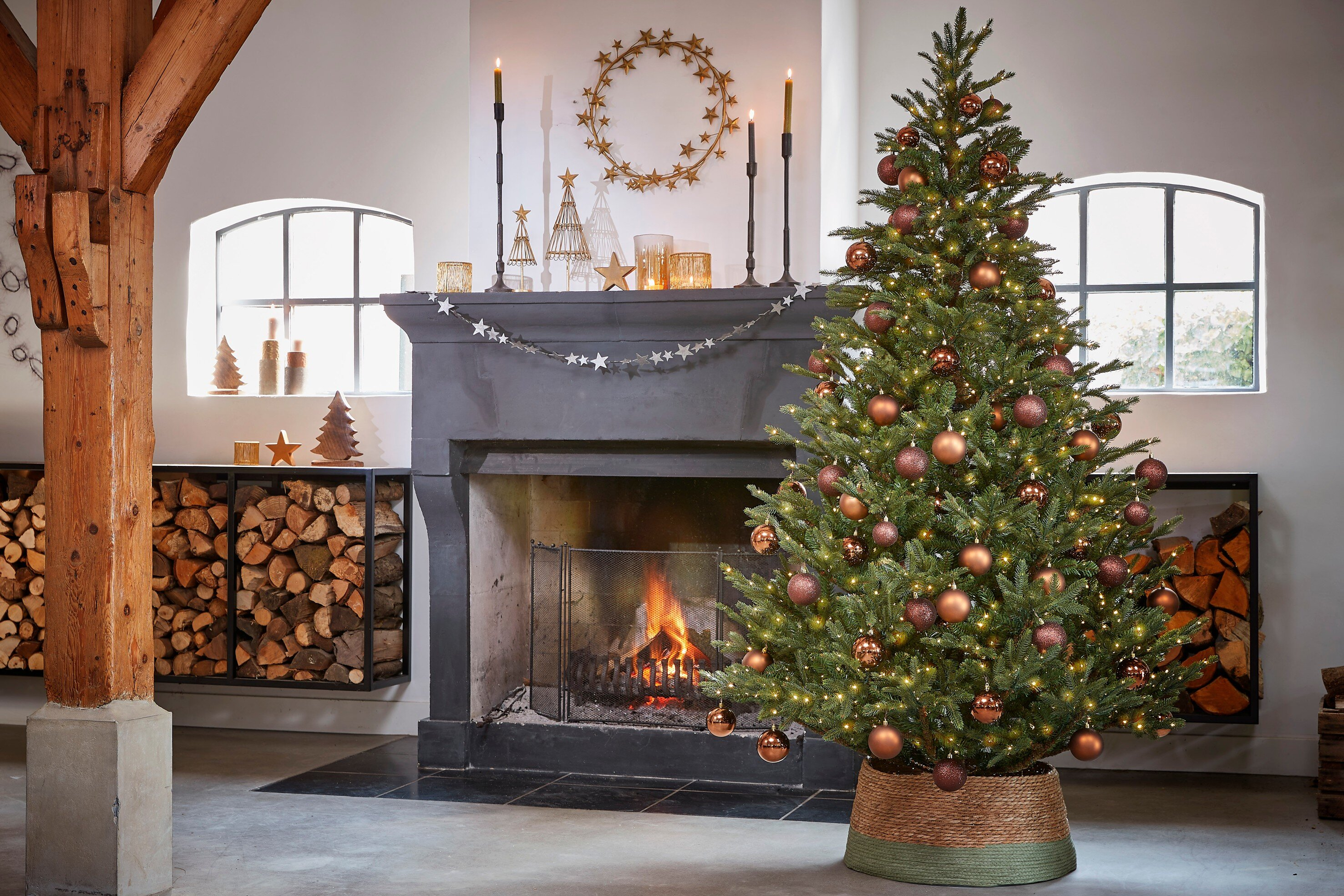 Un sapin de Noël décoré près d'une cheminée avec du feu. Une poutre en bois à gauche.