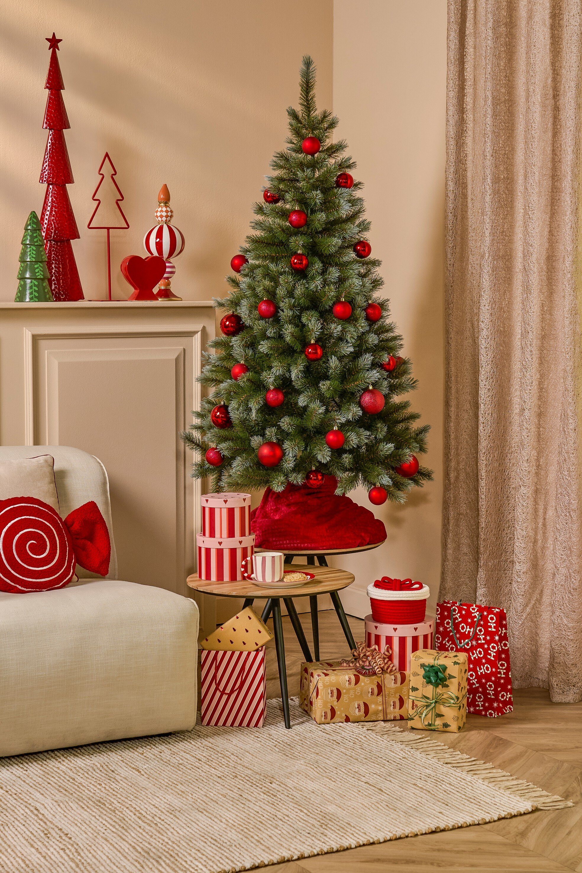 Un sapin de Noël décoré avec des ornements rouges et des cadeaux. Un canapé, un tapis et des décorations sont dans la pièce.