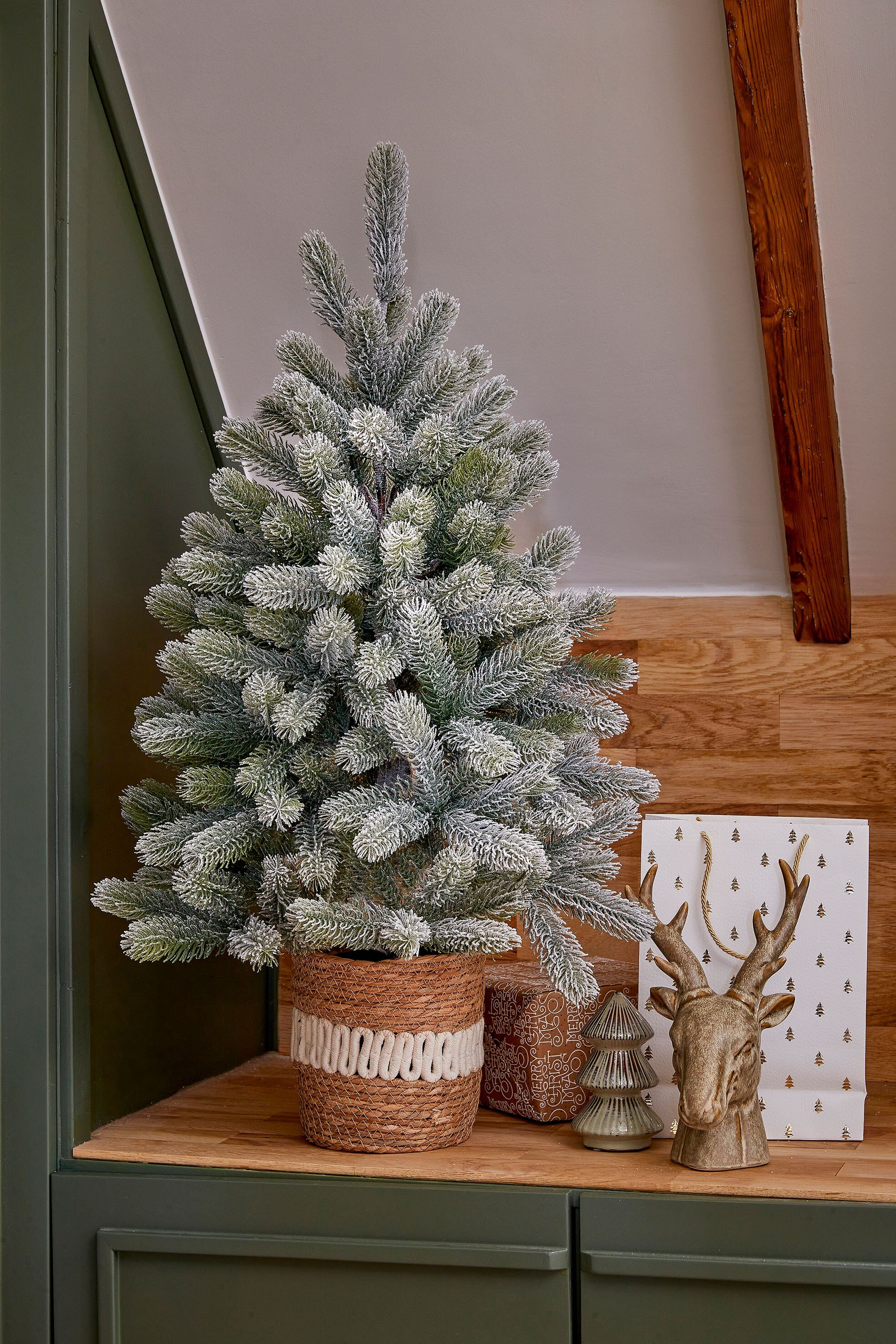 Un petit sapin de Noël dans un pot tissé, avec des décorations dont une tête de cerf et un sac cadeau.