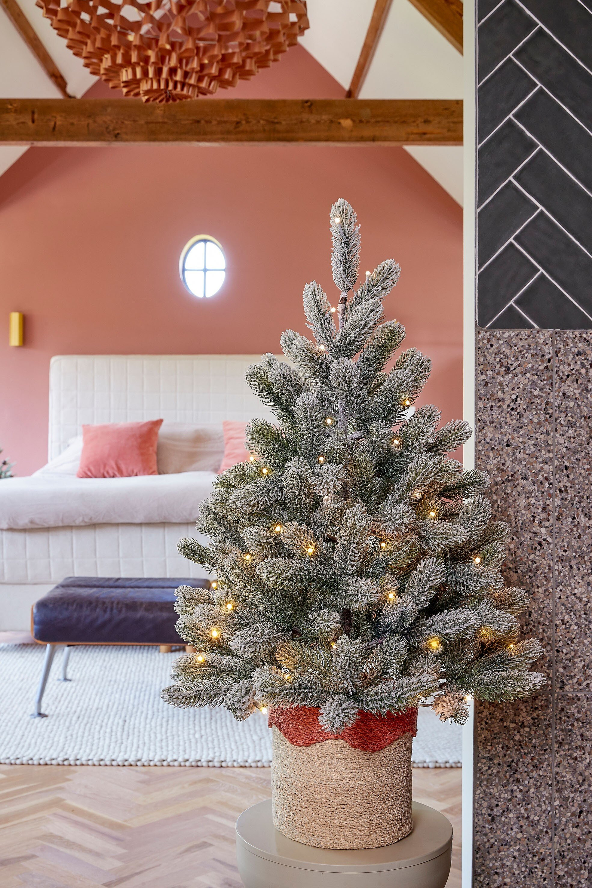 Un sapin de Noël décoré dans un pot, avec des lumières, devant une chambre avec des murs roses et un lit.