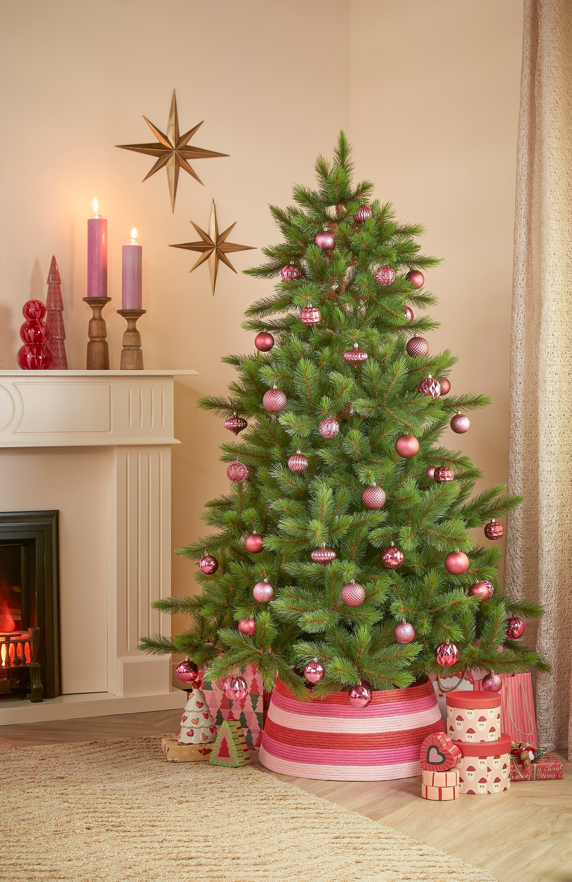 Un sapin de Noël décoré avec des ornements roses. Cheminée et bougies à gauche. Cadeaux et décoration étoilée présents.