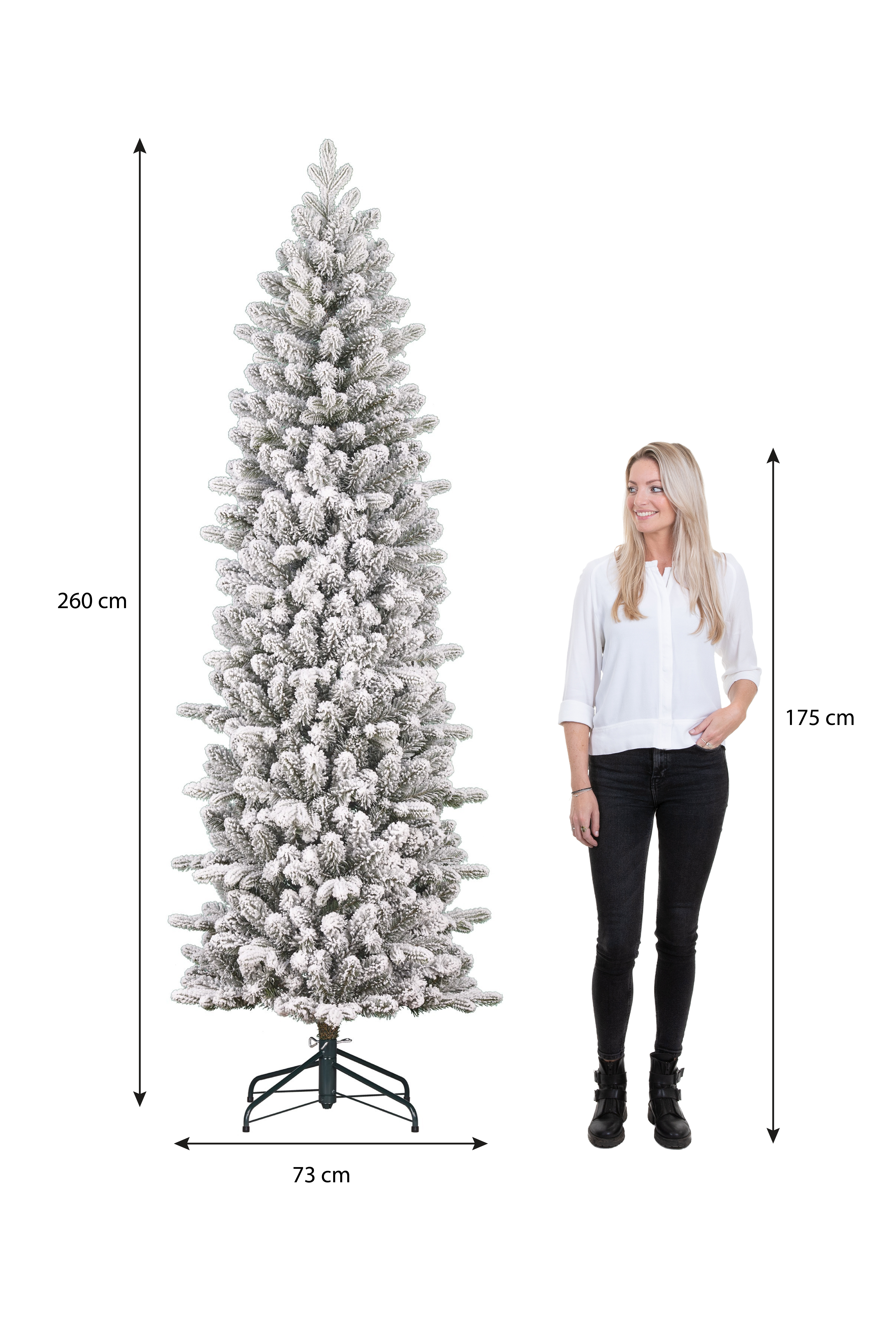 Un grand sapin de Noël enneigé se tient à côté d'une femme en pantalon noir et chemise blanche.