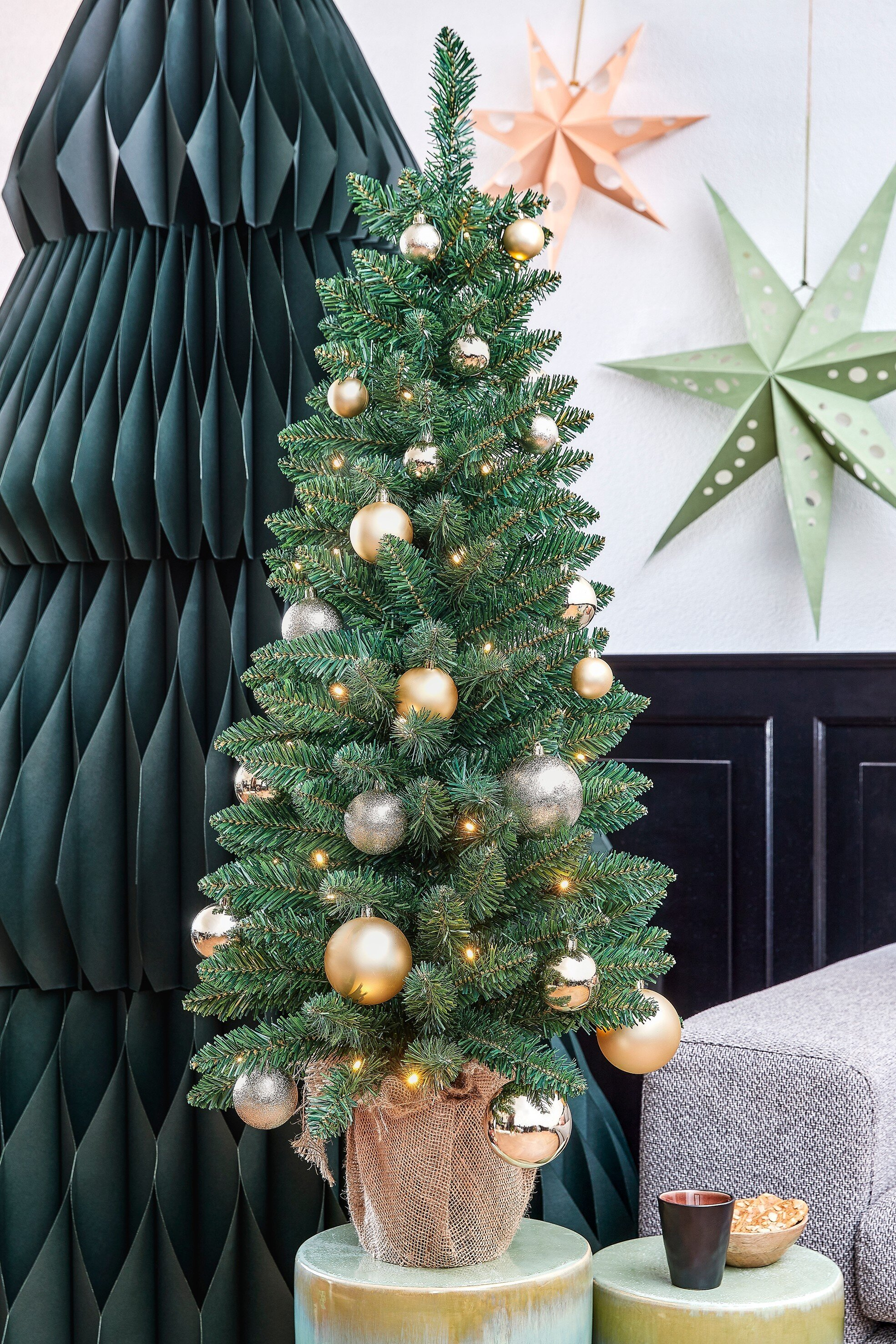 Un sapin de Noël décoré avec des ornements, des lumières et une base en toile de jute, avec des étoiles en papier et un arbre vert foncé en arrière-plan.