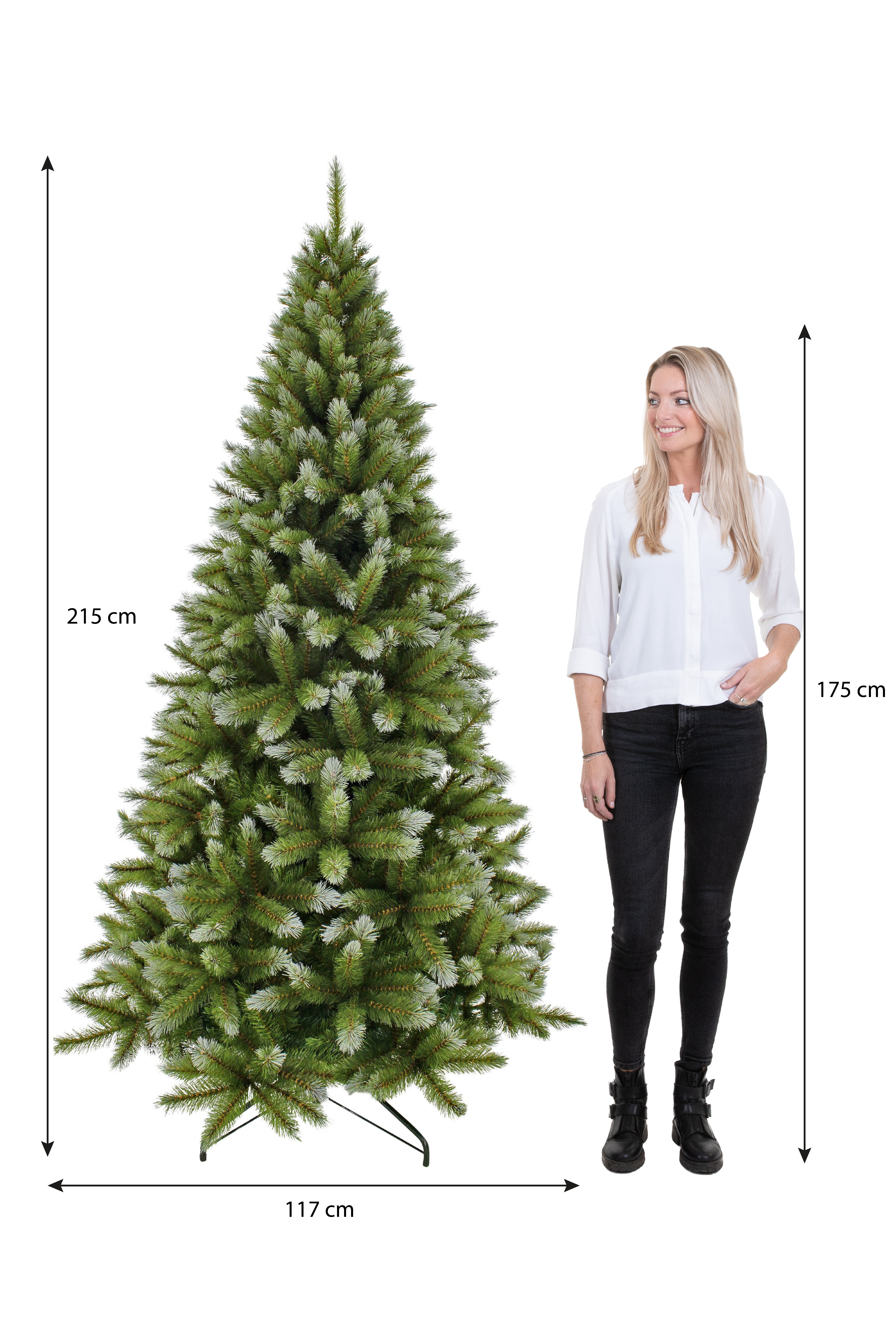 Een vrouw staat naast een grote kerstboom. De boom is groen met witte uiteinden. De vrouw draagt een witte top en een zwarte broek.