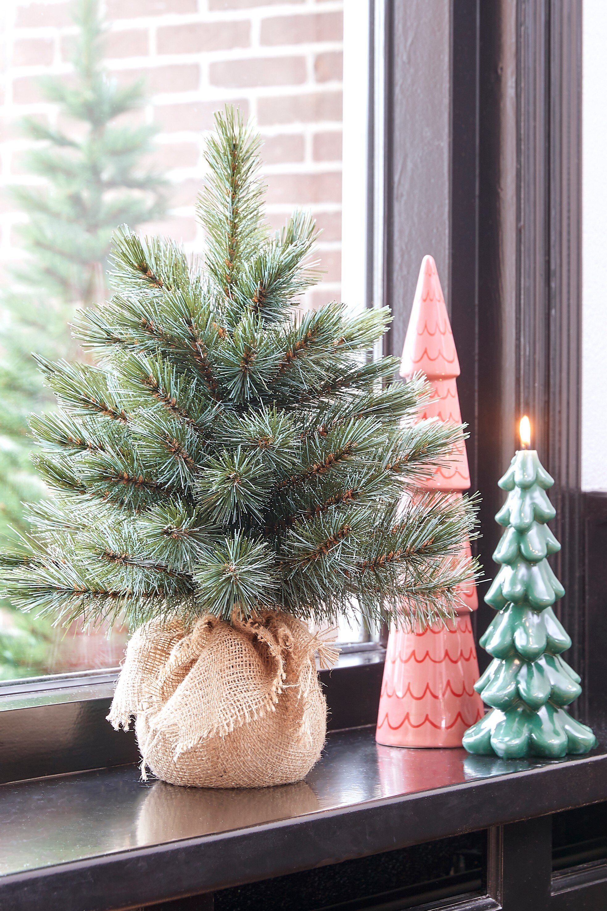 Un petit arbre de Noël dans un sac en toile de jute, à côté d'un arbre en céramique rose et d'une bougie.