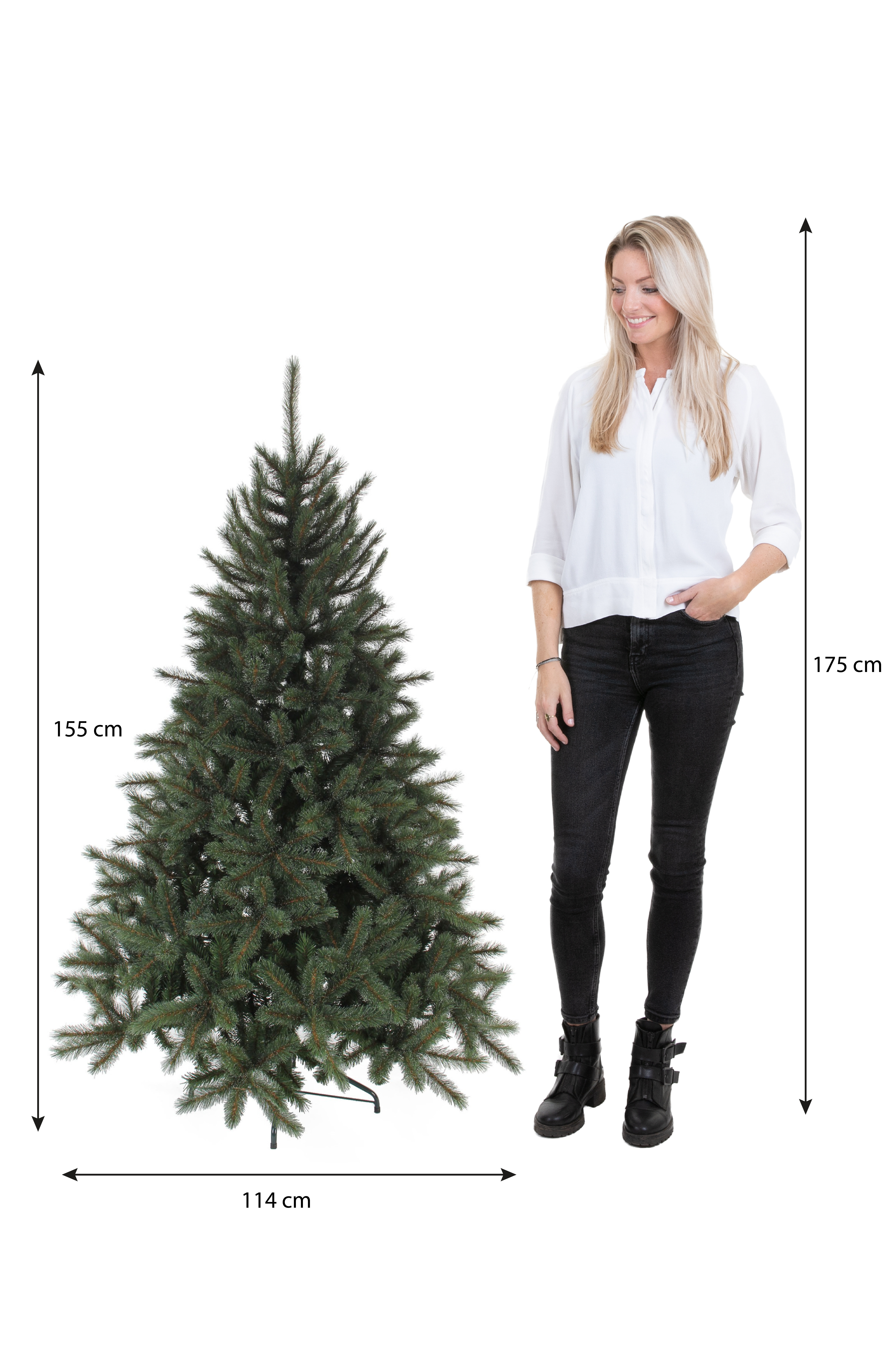 Une femme se tient près d'un sapin de Noël. L'arbre est vert. La femme porte une chemise blanche et un jean noir.